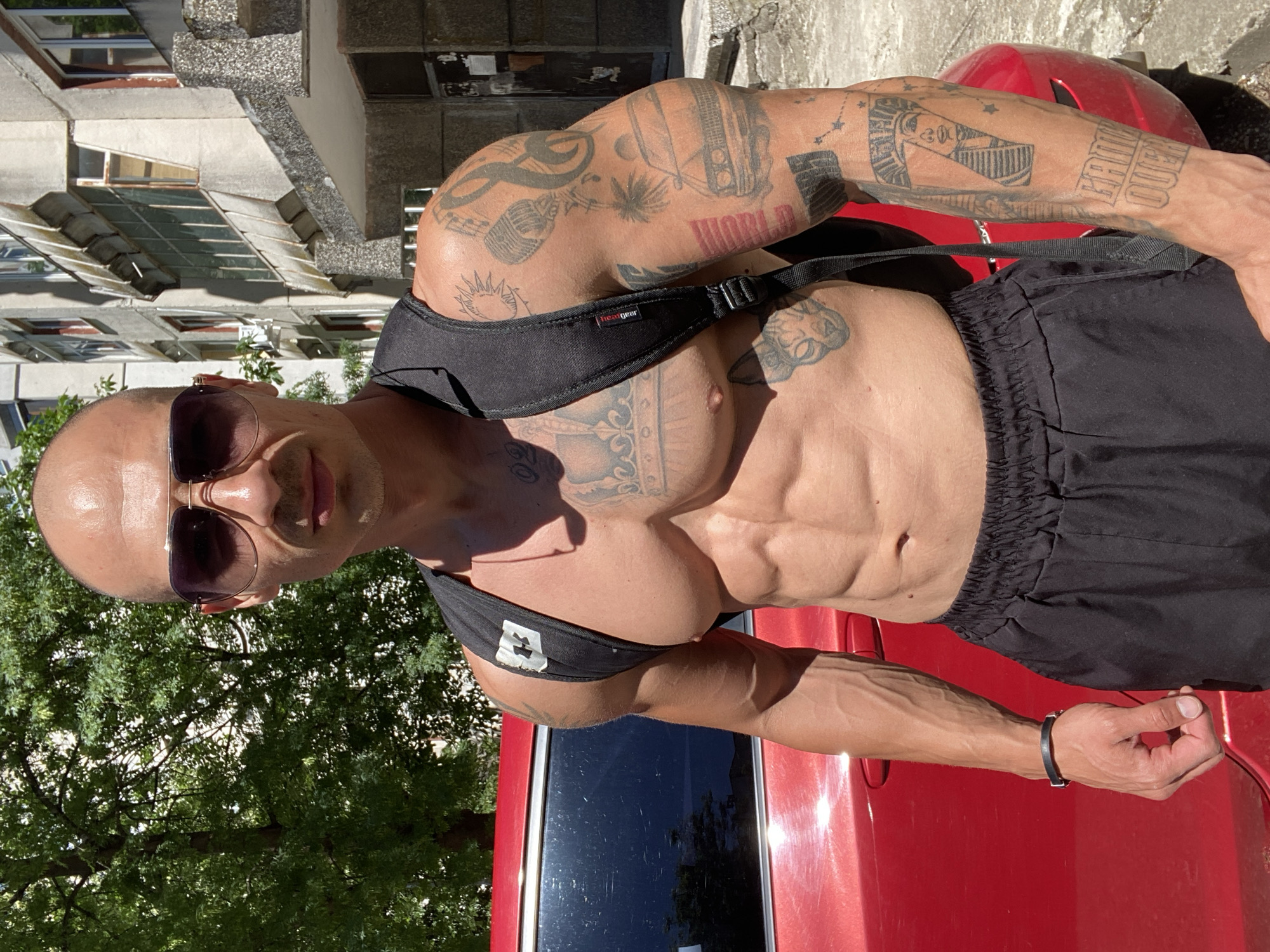 The image shows a muscular, bald man standing outdoors in front of a red car and a building. The man is wearing sunglasses and a black tank top, with a black backpack. His arms and chest are covered in tattoos. The building in the background appears to be an apartment building with multiple windows and balconies. The overall scene suggests a casual setting, possibly on a street or in a parking lot in Pleven, Bulgaria. The lighting indicates it is daytime, with shadows cast by the sun.