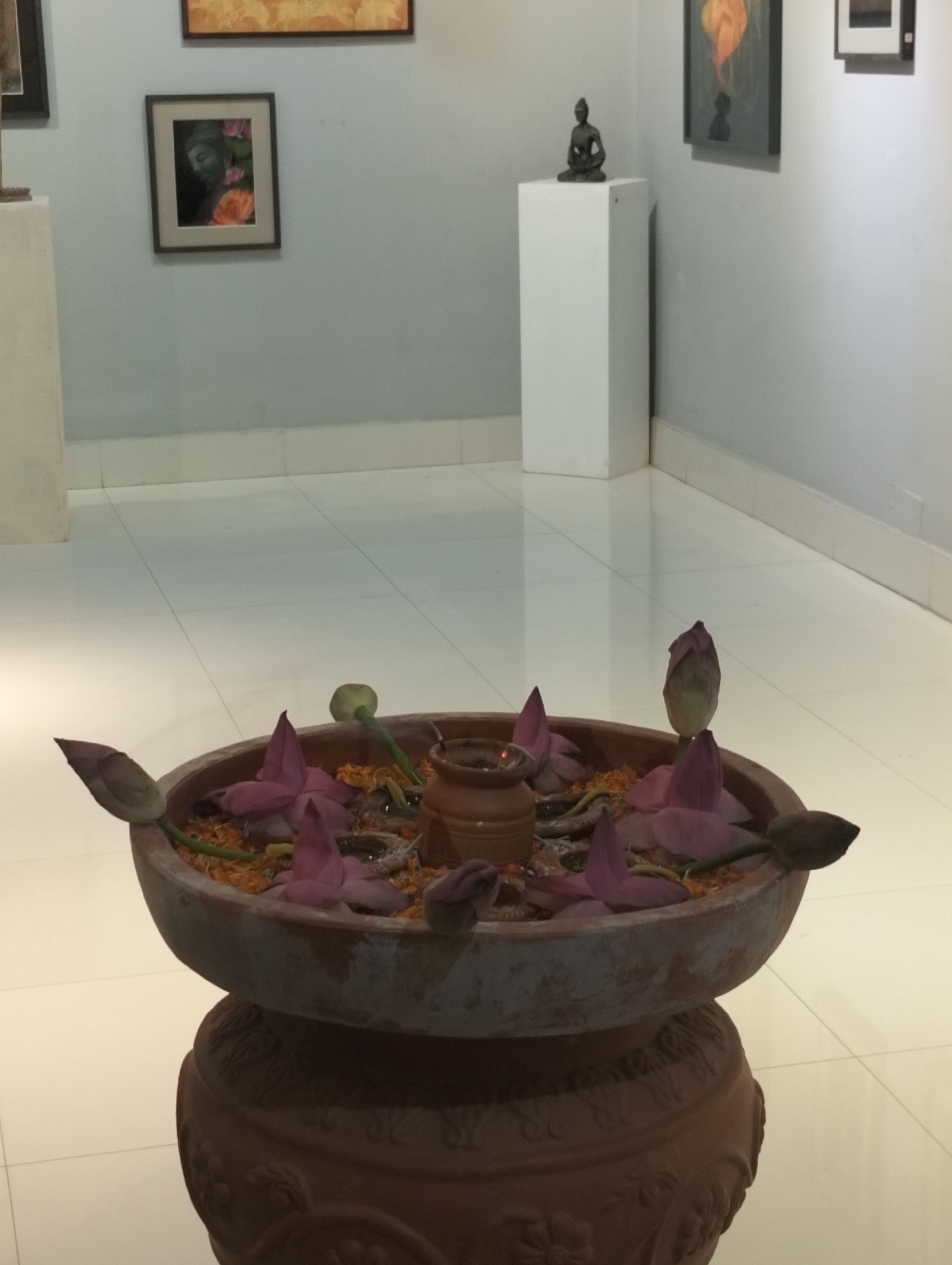 The image depicts an indoor setting, likely an art gallery, in Dhaka, Bangladesh. The foreground is dominated by a large, weathered, terracotta-colored planter bowl resting on a matching, ornately decorated pedestal. The bowl contains water and a variety of lotus flowers, both open and closed buds, along with loose orange petals scattered among them.
In the background, a series of art pieces are displayed on the walls. To the left, a framed image shows a depiction of Buddha intertwined with floral imagery. Further to the right, a white plinth supports a small statue of Buddha. Additional framed artworks are visible on the walls, including one with an orange and gray color scheme. The walls are off-white, and the floor is covered with light-colored, square tiles. The overall lighting is diffused and bright, providing adequate illumination to the gallery space.