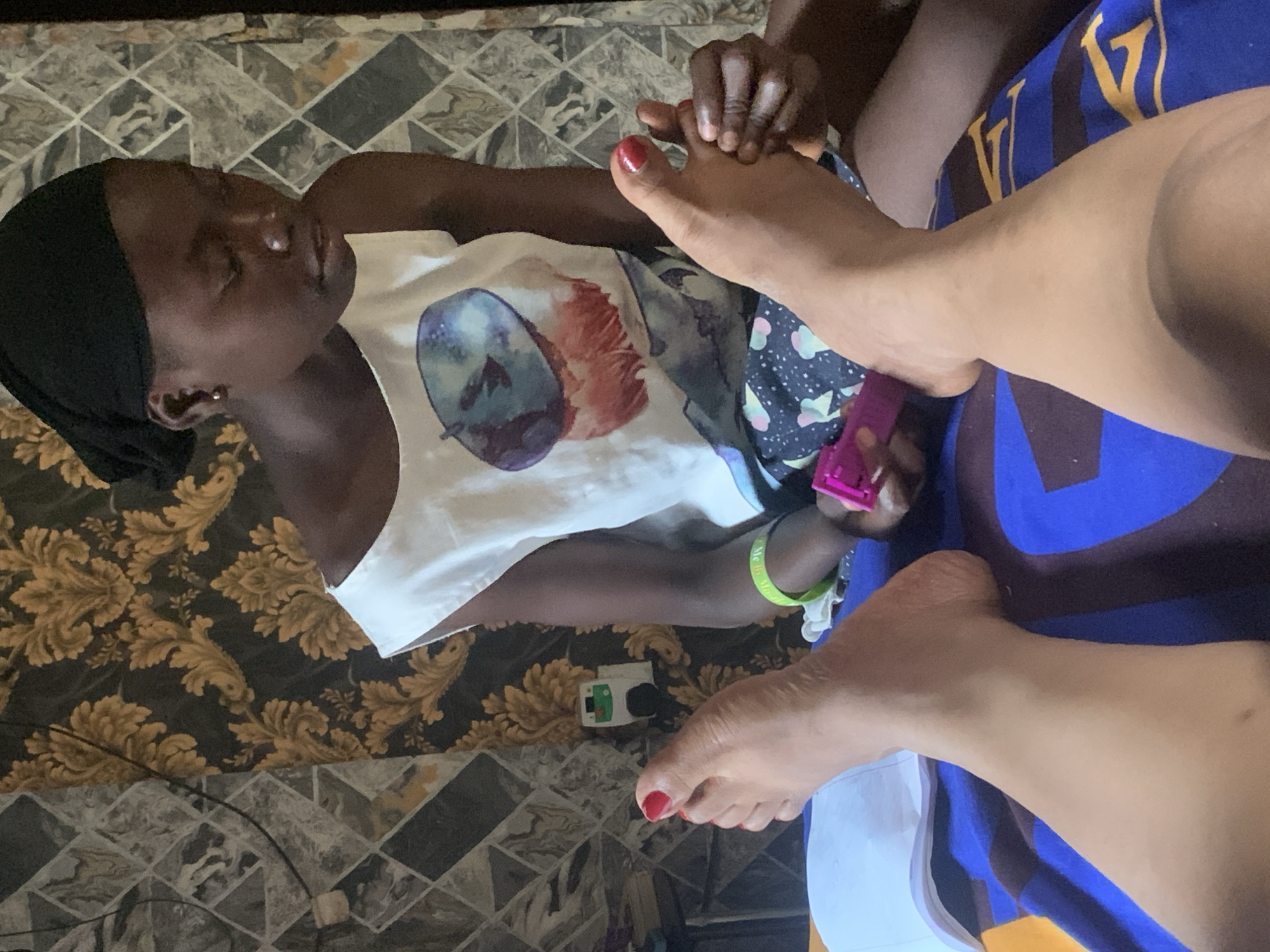The image captures an overhead view of two individuals engaged in a foot massage in what appears to be an indoor setting. One individual, with dark skin and wearing a black headscarf, lies down, with their feet positioned towards the viewer. They are wearing a white sleeveless top with a colorful abstract print. The other individual, whose legs and arms are visible, is massaging the feet of the first person. This person is wearing a blue garment with yellow lettering that appears to be a logo.
The scene is set on a floor covered with patterned tiles, with a large, ornate floral rug to the left. A small white electrical device with green and red accents is visible on the tiled floor. The lighting suggests it is daytime. The activity appears to be relaxed and intimate, possibly a self-care or pampering session. There is no visible text that provides specific context beyond what is on the clothing. The location is identified as Sabon Tasha, Nigeria.