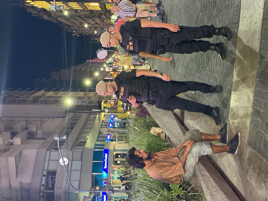 Two uniformed officers stand near a man sitting on a ledge. The officers are wearing dark uniforms and light berets. The seated man, with long dark hair and a beard, wears a tan shirt and gray shorts. They are on a paved area at night with buildings and street lights in the background. The scene is lit, revealing details of the surroundings.