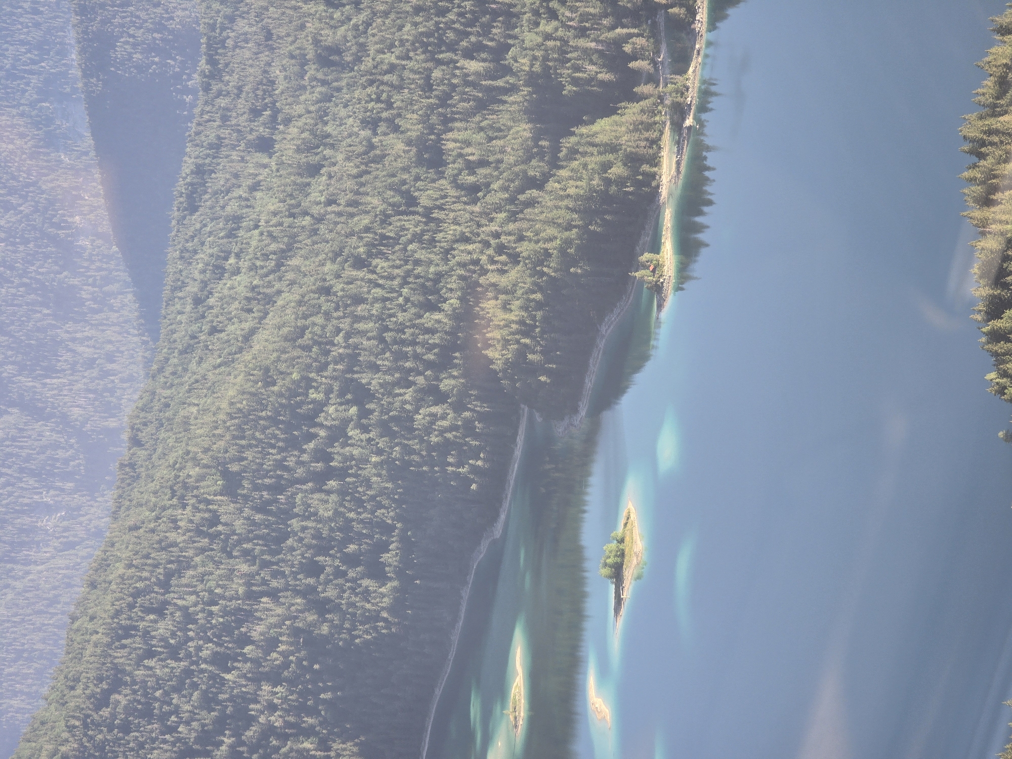 The image is an aerial view of a vast, pristine alpine lake in Grainau, Germany, likely Eibsee, surrounded by dense evergreen forests that climb the steep slopes of the surrounding mountains. The lake's water is a striking turquoise and deep blue, with areas of lighter turquoise indicating shallower regions or sandbars. Two small islands, each covered in lush green trees, dot the lake. One island is closer to the foreground and appears to have a small, red-roofed structure. The other island is further in the distance. The forested mountains are layered, with the closest ones covered in a thick carpet of trees, while those in the background are softer, more muted in color due to atmospheric haze. Sunlight casts a gentle glow on the scene, highlighting the textures of the trees and the shimmering surface of the water. No people or man-made structures other than the small building on the island are visible.