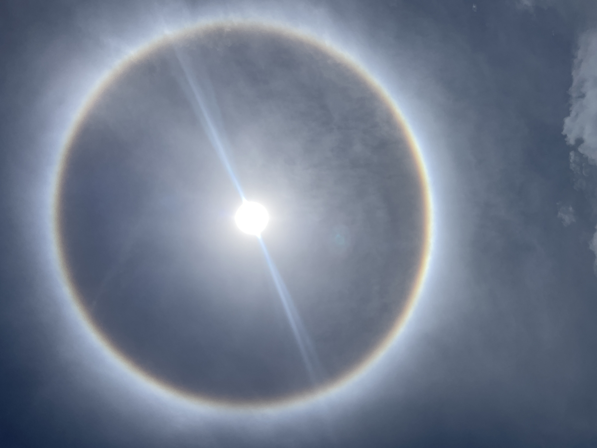 The image shows a solar halo, a ring of light appearing around the sun, in the sky. The sun is in the center of the halo, and a bright streak of light extends downwards from it. The sky is overcast with thin, patchy clouds. The exact location in the city of Altagracia, Venezuela is not identifiable from the image. There are no people or other identifiable objects in the image.