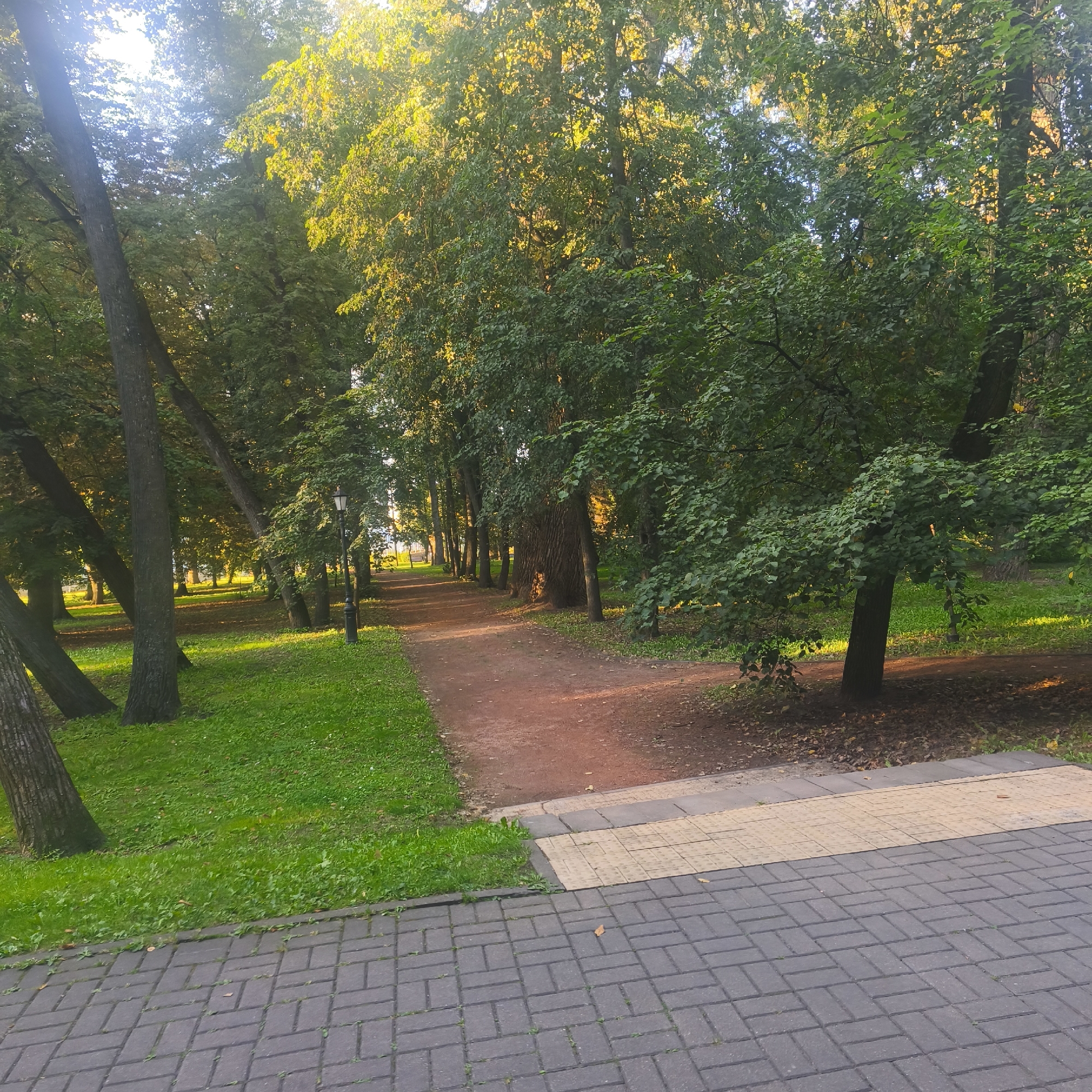 The image depicts a park pathway lined with trees on either side. The pathway is made of reddish-brown dirt, and it curves into the distance. To the left of the pathway, there is a grassy area with several large trees. To the right, the pathway is bordered by a strip of yellow tactile paving, followed by a gray brick-paved walkway. Sunlight filters through the trees, creating dappled shadows on the ground. The scene suggests a tranquil setting within a park. This location is likely in the city of Minsk, Belarus, given the presence of similar park layouts and the context of the request. There are no people or animals visible in the image.