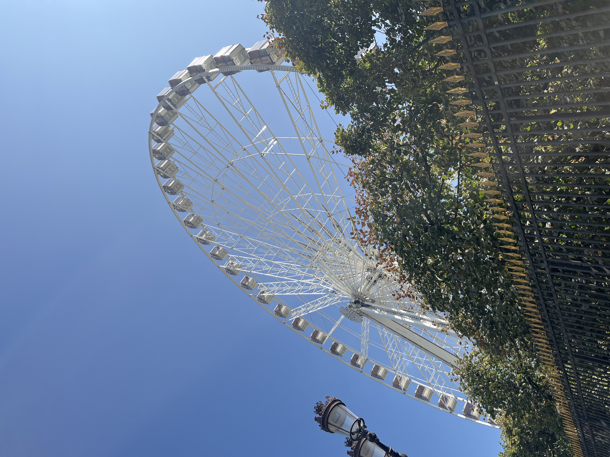 The image depicts a close-up, low-angle view of a large Ferris wheel against a clear blue sky. The Ferris wheel is white with numerous gondolas. In the foreground, a black wrought-iron fence with pointed gold finials runs horizontally across the bottom of the frame. Above the fence, green trees with some autumnal foliage are visible. To the left of the Ferris wheel, a decorative street lamp with two lights can be seen. This scene is consistent with the location of the Rueil-Malmaison Ferris Wheel in Paris, France, a temporary attraction that appears in this image. No people are visible in the frame.