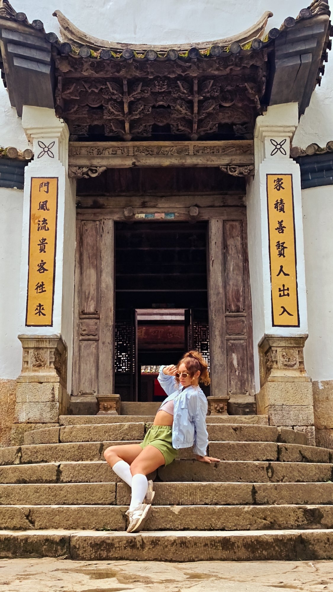 A young woman is sitting on stone steps in front of a traditional building in Sà Phìn Commune, Vietnam. The building has an elaborate wooden facade with Chinese characters on the pillars. The woman is wearing sunglasses, a light blue jacket, green shorts, white socks, and sneakers. She is looking up and holding her hand up to shield her eyes from the sun. The stone steps lead up to the entrance of the building.