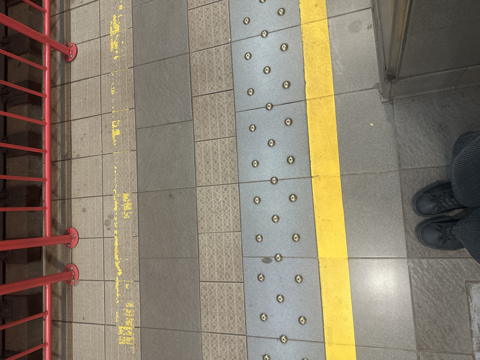 A high-angle, medium shot shows a section of a subway platform in Sofia, Bulgaria, with a yellow safety line demarcating the edge. In the foreground, two black leather sneakers and the bottom of dark corduroy pants are visible, suggesting a person is standing on the platform. The platform is tiled with various patterns and colors, including grey tiles with raised circular indicators and a textured pattern. Behind the yellow line, more grey tiles are visible. Further back, red metal railings stand over train tracks. The lighting is artificial and casts some shadows.