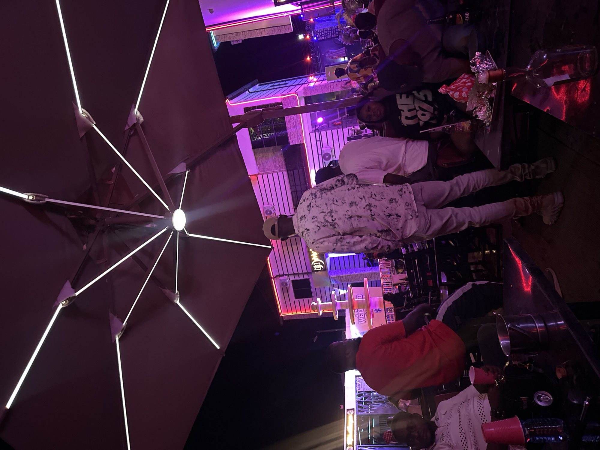 The image depicts a night scene, likely outdoors, in an entertainment venue, possibly in Tumfure, Nigeria, though this is not explicitly confirmed visually. Several people are present at tables, engaged in social activities. One person stands, wearing a patterned shirt and light-colored pants. Another individual wears a red shirt. Tables are cluttered with items including cups, bags, and what appears to be an empty bottle. The venue is illuminated with colorful lighting, including purple and blue hues. An umbrella-like structure with integrated lights is visible in the foreground. The overall environment suggests a relaxed, nightlife setting.