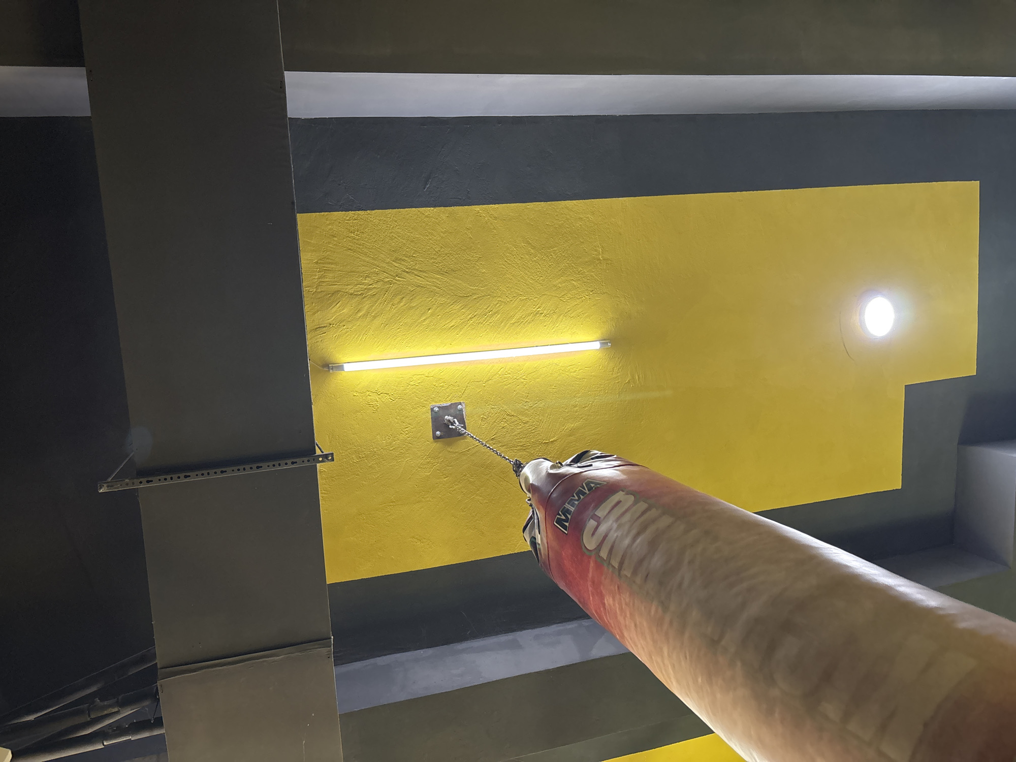 This media file captures a low-angle, upward view of a gym ceiling in Botevgrad, Bulgaria. The dominant feature is a punching bag, a thick, cylindrical object with red and beige coloring, hanging from a chain attached to a metal fixture on the ceiling. The bag has the letters "MMA" visible on its side.
The ceiling itself is painted in distinct sections. A large area is a bright, textured yellow, contrasting with darker grey sections. A long, linear fluorescent light fixture is mounted on the yellow portion, illuminating the space. Another, smaller circular light fixture is also visible on the yellow ceiling. Structural elements, such as dark grey beams and what appear to be air ducts or conduits, are visible in the darker grey sections of the ceiling. One dark grey beam has a metal bracket attached to it, with a series of holes.
The overall impression is of an indoor gym environment, likely a boxing or mixed martial arts facility, given the presence of the punching bag. The lighting suggests it could be daytime, but the artificial light is the primary illumination source. There are no people visible, and the focus is on the equipment and the architectural elements of the ceiling. The image conveys a sense of the functional, perhaps slightly industrial, aesthetic common in such training spaces.