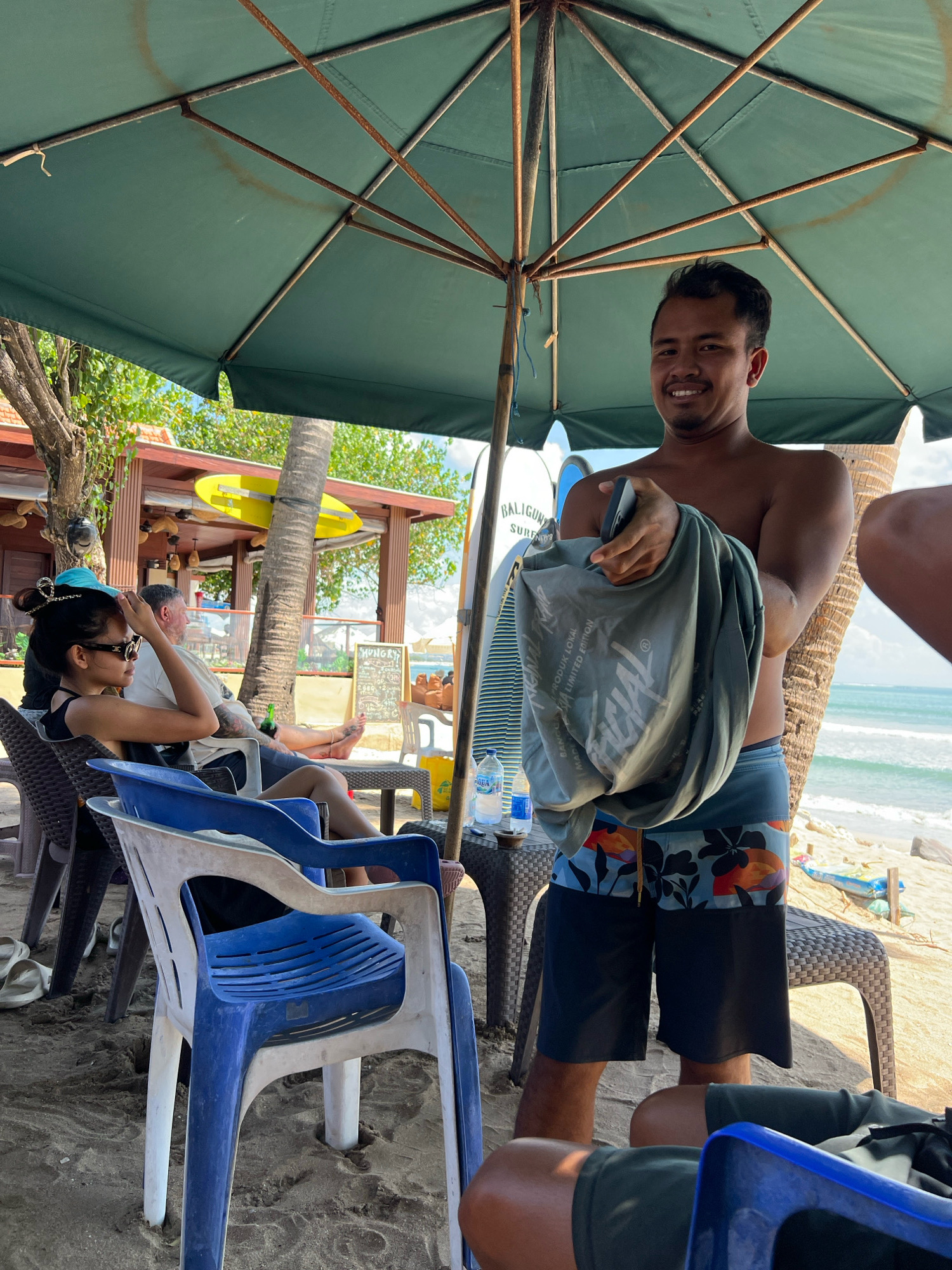 A man in dark floral swim trunks and no shirt stands under a large green beach umbrella, holding a gray t-shirt. He is smiling at the camera. Behind him, a woman wearing a black tank top and sunglasses sits in a chair, adjusting her hat. Next to her, a man in a white t-shirt reclines. In the background, surfboards lean against a palm tree, and the ocean waves crash on the sandy beach. The scene appears to be in Kuta, Bali, Indonesia, a popular tourist destination known for its beaches and surf spots.