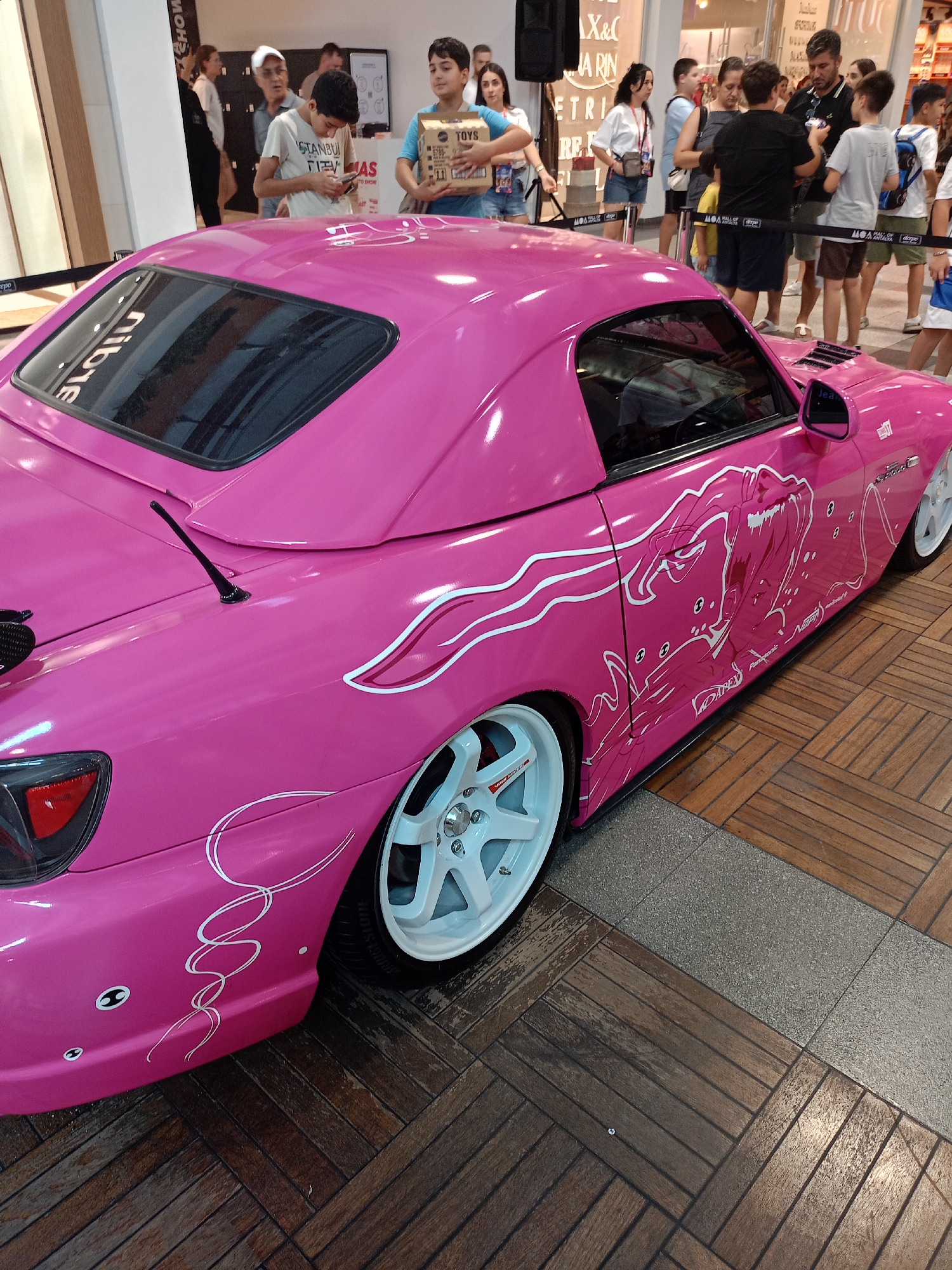 The image shows a bright pink Honda S2000 with white anime-style graphics on its side, parked indoors on a wooden floor. Several people are visible in the background, including men, women, and children, some of whom are looking at the car. The setting appears to be a shopping mall, possibly the Mall of Antalya in Kepez, Turkey, as indicated by visible signage. The car is a prominent focal point, with its custom paint job and large white wheels.