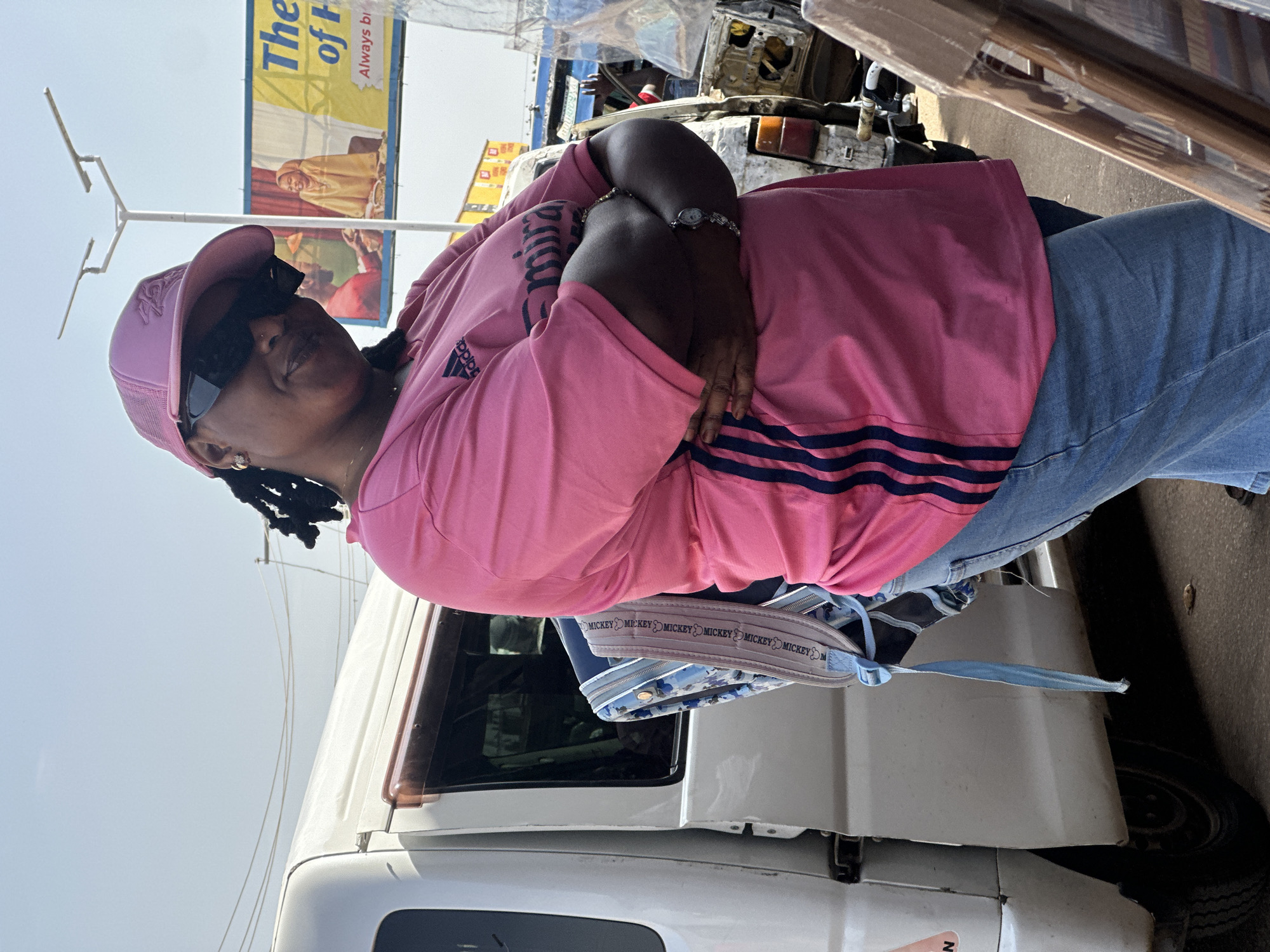A full-bodied Black woman in a pink Adidas t-shirt, blue jeans, and a pink baseball cap stands with her arms crossed in front of a white van in Bukuru, Nigeria. She wears black sunglasses and a silver wristwatch. A blue backpack with a Mickey Mouse pattern is slung over her shoulder. In the background, a billboard advertises "The ... of H" with the words "Always b". The scene is outdoors, with a clear sky and power lines visible. The ground is paved.