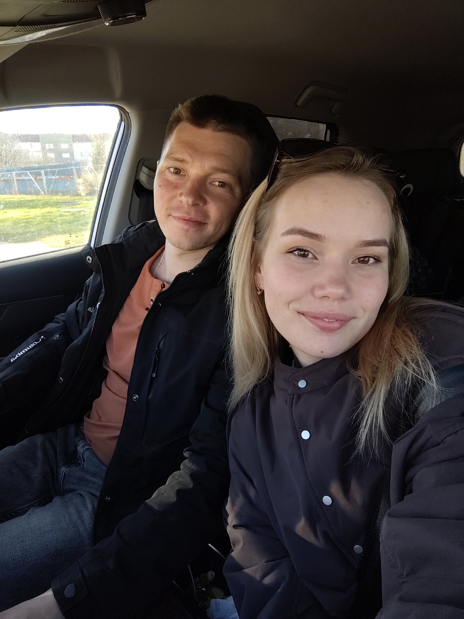 A medium-close-up, eye-level selfie captures a man and a woman seated in the front passenger and driver's seats of a car, respectively. The setting is bright daylight, suggesting a sunny day, with the sun casting highlights through the car's side window. The background visible through the window shows a grassy area, trees, a blue fence, and some distant buildings, indicating a rural or suburban environment in Марковский, Russia.
The man, positioned on the left side of the frame, has short dark hair and brown eyes, looking towards the camera with a subtle smile. He is wearing a dark jacket over an orange collared shirt, with a silver chain visible around his neck. His left arm is resting on the car's interior. Partially visible text on his jacket sleeve appears to be "Adium" or a similar brand name.
The woman, on the right, has blonde hair, brown eyes, and a pleasant, gentle smile directed at the camera. She is wearing a dark, possibly padded, jacket with silver snap buttons down the front. Sunglasses are resting on top of her head. Both individuals appear relaxed and content. The car's interior, including the roof lining and a handle, is visible above them.