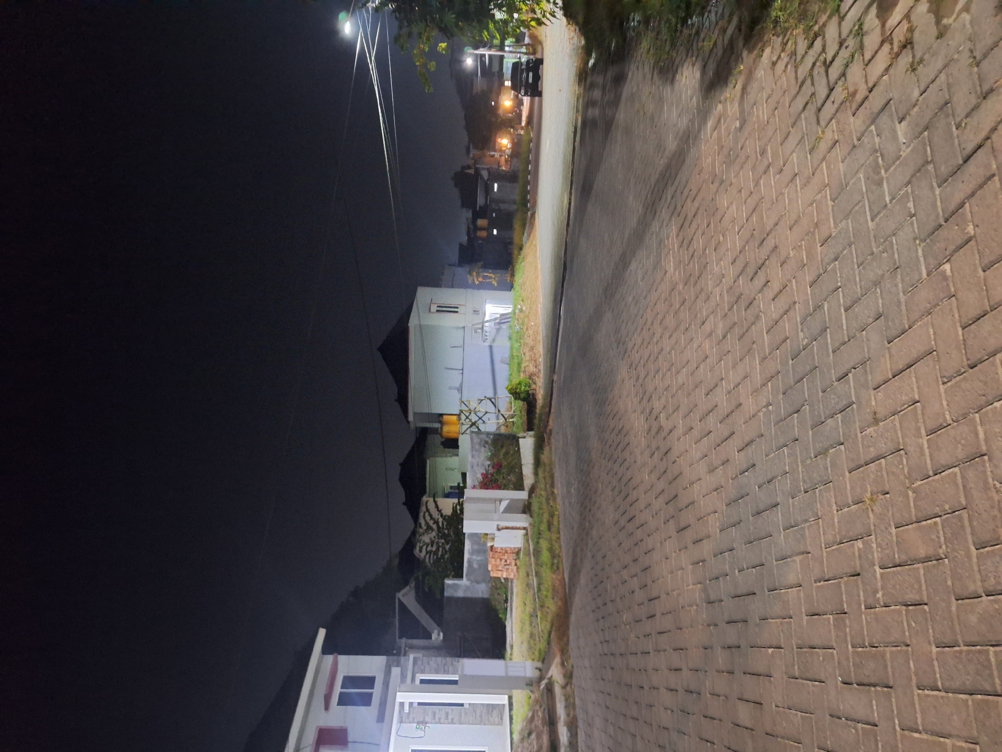 An outdoor, nighttime, vertical, medium shot captures a paved walkway in the foreground, leading towards a street and several residential buildings in the background. The walkway is constructed of reddish-brown rectangular pavers laid in a herringbone pattern. On the left, a modern, two-story house is visible, primarily white with red accents around the upper windows. To the right of this house, a grassy area separates it from a low, grey stone wall. Beyond the wall, more houses with dark roofs are situated, some with illuminated windows and outdoor lighting. A single black SUV is parked on the street in the middle ground. Streetlights and overhead wires are present, contributing to the ambient illumination of the scene. The sky is dark, indicating nighttime. The overall setting suggests a residential neighborhood.