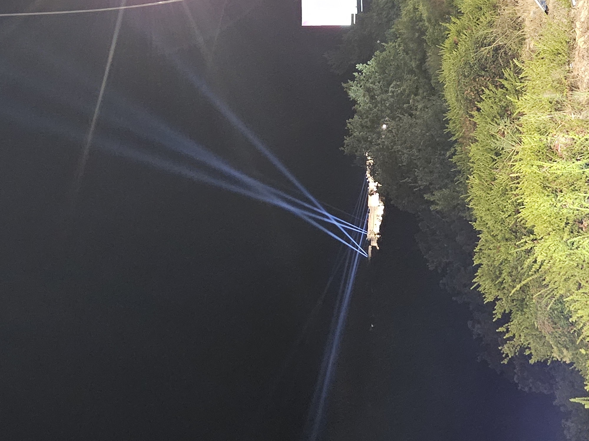 In an outdoor nighttime scene, several powerful white spotlights pierce the dark sky, creating visible beams that converge on a distant, illuminated structure. The structure appears to be on elevated ground, with foliage in the foreground and midground obscuring parts of the landscape. The spotlights are angled upwards and outwards, suggesting a directed light show or illumination event. The sky is a deep black, with no visible celestial bodies. The foreground is dominated by dense green vegetation, which appears to be a hillside covered in bushes and smaller trees. The overall impression is one of a dramatic outdoor event at night.