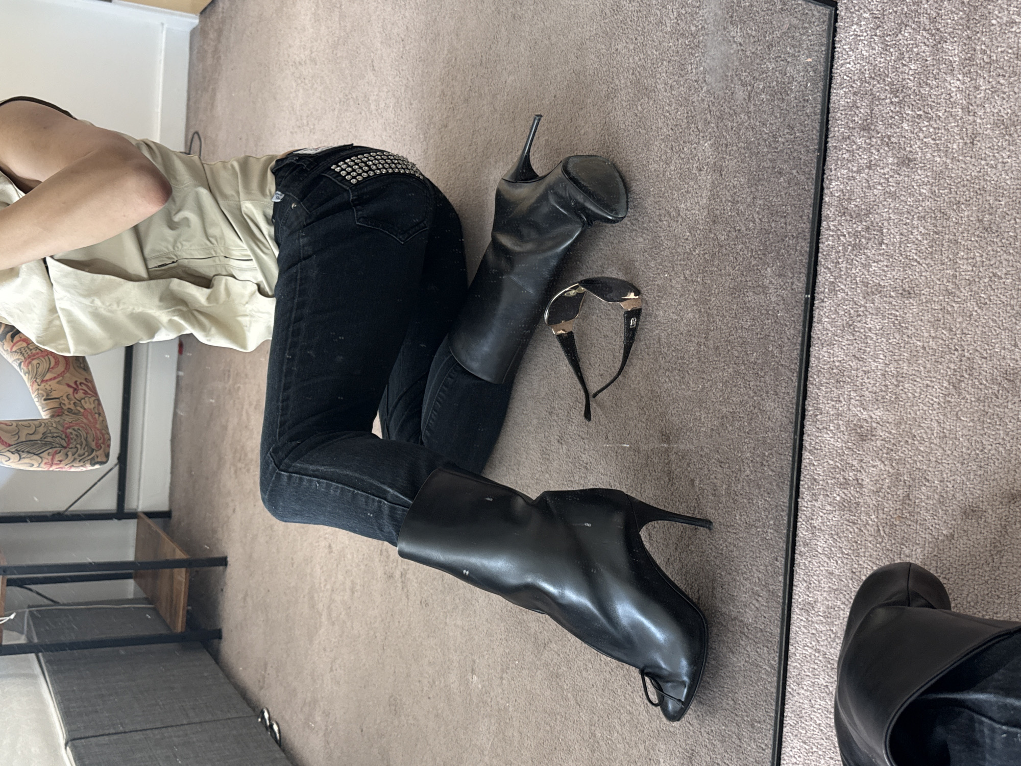 The image captures a person's reflection in a large mirror that is placed on a light brown or grey carpeted floor. The person is seen lying horizontally within the frame, showcasing their outfit and accessories. They are wearing fitted black jeans, which feature a prominent studded design on the back pocket. Their upper body is partially visible, clothed in a light-colored (possibly beige or off-white) collared shirt or jacket. One arm, covered in intricate red and black tattoos, is also visible.
One black high-heeled ankle boot is worn on the person's foot, while its matching pair lies unzipped on the carpet next to them. This boot has a distinctive design, including a small toe-loop detail. A pair of black sunglasses with silver accents is also casually placed on the carpet near the boots.
The reflection in the mirror reveals parts of an indoor setting, including a white wall, a black metal shelving unit with a wooden plank, and a piece of grey upholstered furniture, possibly a sofa or bench. The black edge of the mirror is visible, separating the direct view of the carpet from the reflected scene. The scene suggests an outfit-of-the-day or style photo taken indoors, likely in London, United Kingdom, given the provided context. The time of day is not evident. No legible text is visible on any items.
