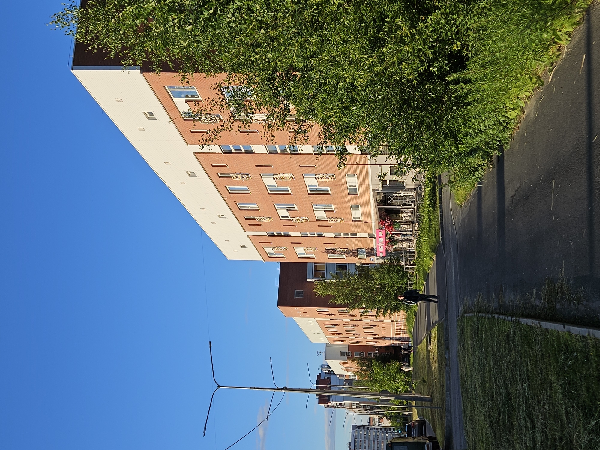 A low-angle, wide shot captures a street scene on a clear, sunny day. The camera is oriented vertically, giving the impression of looking up at the sky, which occupies the left side of the frame. Dominating the right side are two large, multi-story apartment buildings with brick facades and white sections. Lush green trees and bushes flank the buildings and the asphalt road.
In the foreground, a person walks along the sidewalk next to the road. The scene appears to be set in a residential area, likely a city or town, with utility poles and streetlights visible in the distance. The sunlight suggests it's either morning or late afternoon. There is no discernible text in the image.