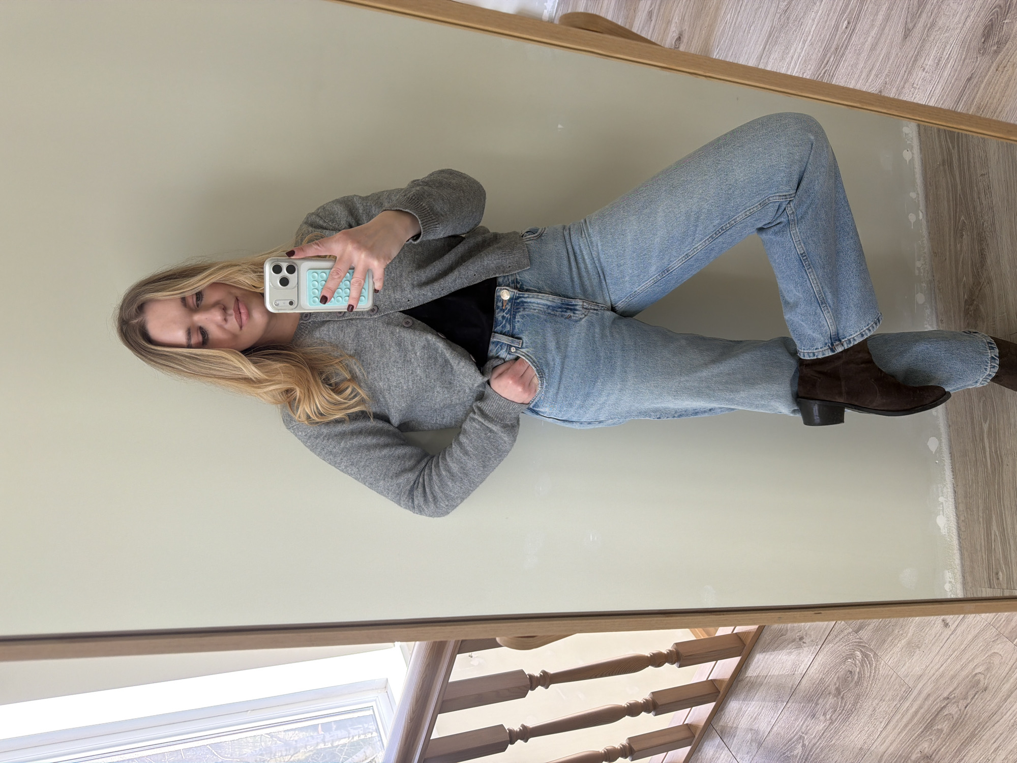 A woman is reflected in a mirror. She is wearing blue jeans, a black shirt, a grey cardigan, and brown boots. She is holding a cell phone with a light blue case in her right hand. The background includes a light-colored wall and a wooden staircase. The location is not specified or discernible from the image to be Dreiliņi, Latvia.