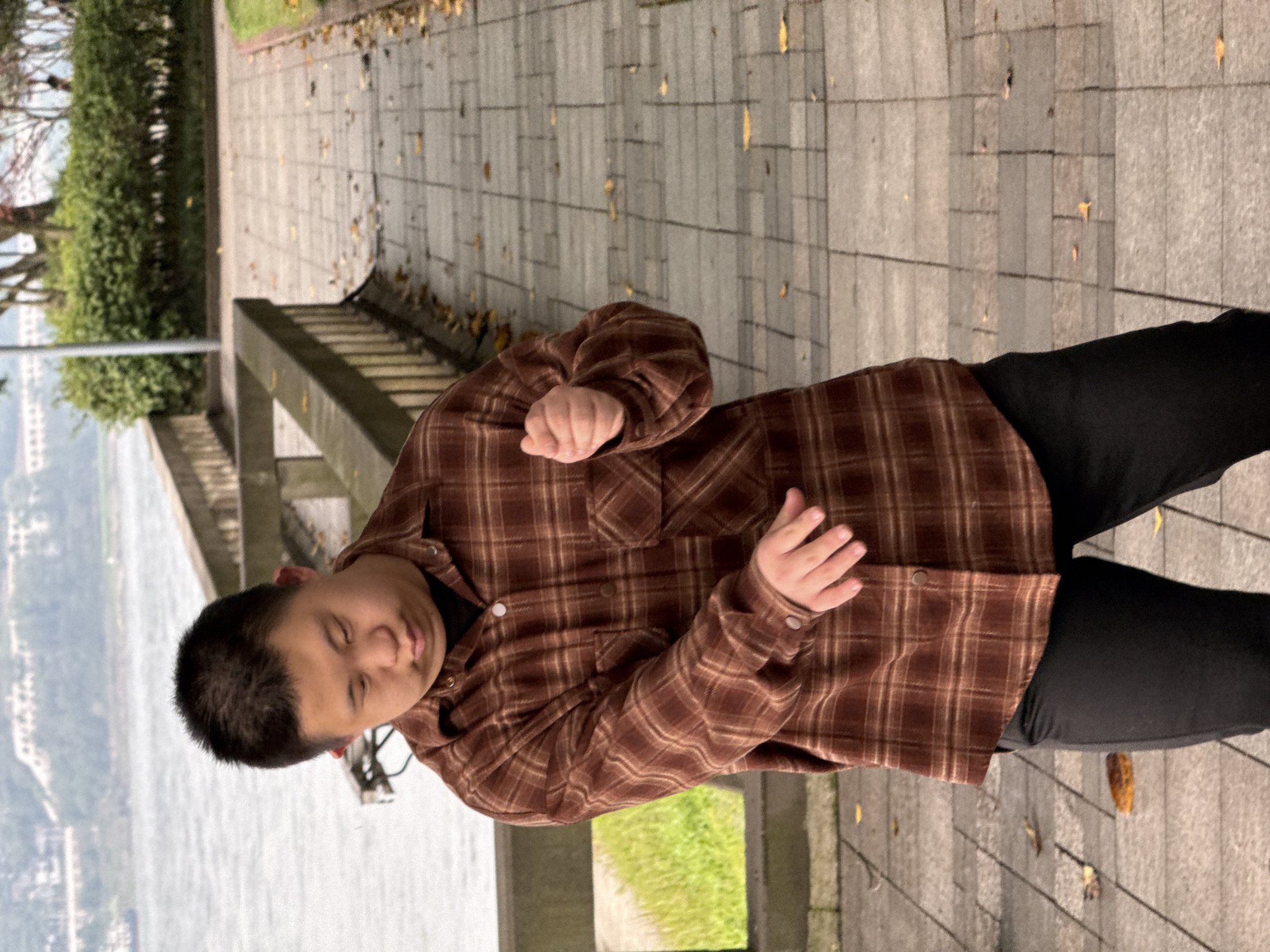 A person is standing on a brick pathway in Jiangjin, China. The person is wearing a brown plaid shirt and black pants. Their hands are outstretched, one in a fist and the other open. The person is looking down with their eyes closed. In the background, there is a body of water, possibly a river, with buildings visible on the opposite bank. There is also a railing beside the pathway, with greenery behind it. The weather appears to be overcast.