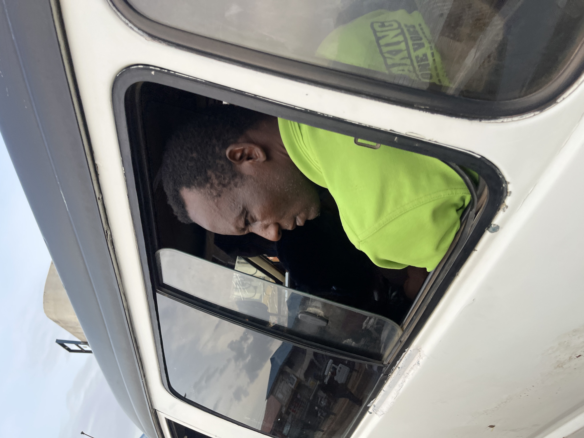 A man wearing a bright green shirt with writing on the back looks out the window of a white van. The van is parked in what appears to be an urban setting. The man's face is in profile, and he has short, dark hair. The interior of the van is dark. The window shows a reflection of buildings and the sky. The man's shirt has the words "MAKING ONE VOICE" printed on the back. The scene is photographed from a slightly elevated angle, looking into the side window of the van. The van's exterior shows some signs of wear and tear.