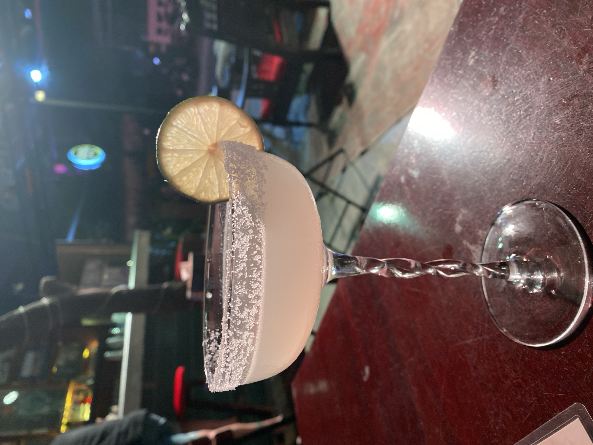 An eye-level shot focuses on a margarita cocktail placed on a dark, distressed wooden table. The glass has a salt-rimmed rim and a twist-stem design, with a lime wedge perched on the edge. The drink itself is a pale, translucent liquid. The background is a blur of activity, suggesting a bar or restaurant setting. In the far background, a person can be seen from the waist down, wearing shorts. To the right, chairs are visible, some with red accents. The lighting is dim, with colored lights creating a vibrant, atmospheric glow, typical of nightlife in Đà Nẵng, Vietnam. The overall impression is that of a relaxed evening out.