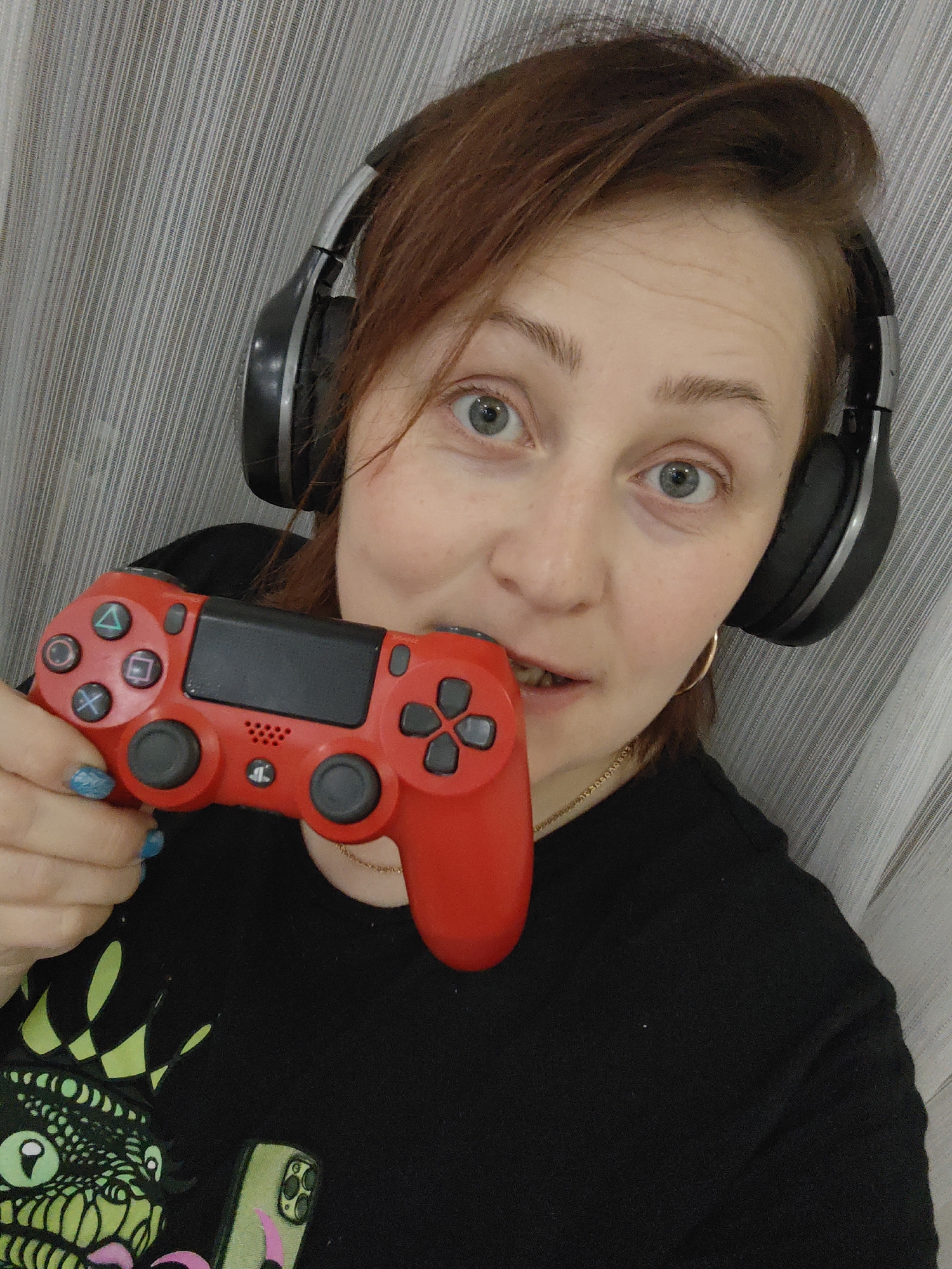 A medium shot captures a young adult female indoors, looking directly at the viewer with wide, light-colored eyes. She has reddish-brown hair and fair skin. She is wearing black over-ear headphones and a black t-shirt with a vibrant green and black graphic design that appears to be a monster or dragon head with a crown. A gold hoop earring is visible on her right ear, and a thin gold chain necklace is around her neck.
She holds a red PlayStation DualShock 4 controller in her left hand, positioned near her mouth, which is slightly open, revealing her teeth, suggesting a moment of intense focus or reaction while gaming. The controller clearly shows the PlayStation symbols (triangle, circle, cross, square) on its face buttons, and the "PS" logo on the home button. A small speaker grille is also visible on the controller. Her left hand has blue nail polish.
In the lower left corner, part of a smartphone in a light green case with a pink design is visible, held near the t-shirt graphic. The background consists of a light-colored, vertically striped curtain or fabric. The overall impression is one of an engaged gamer captured mid-action.
