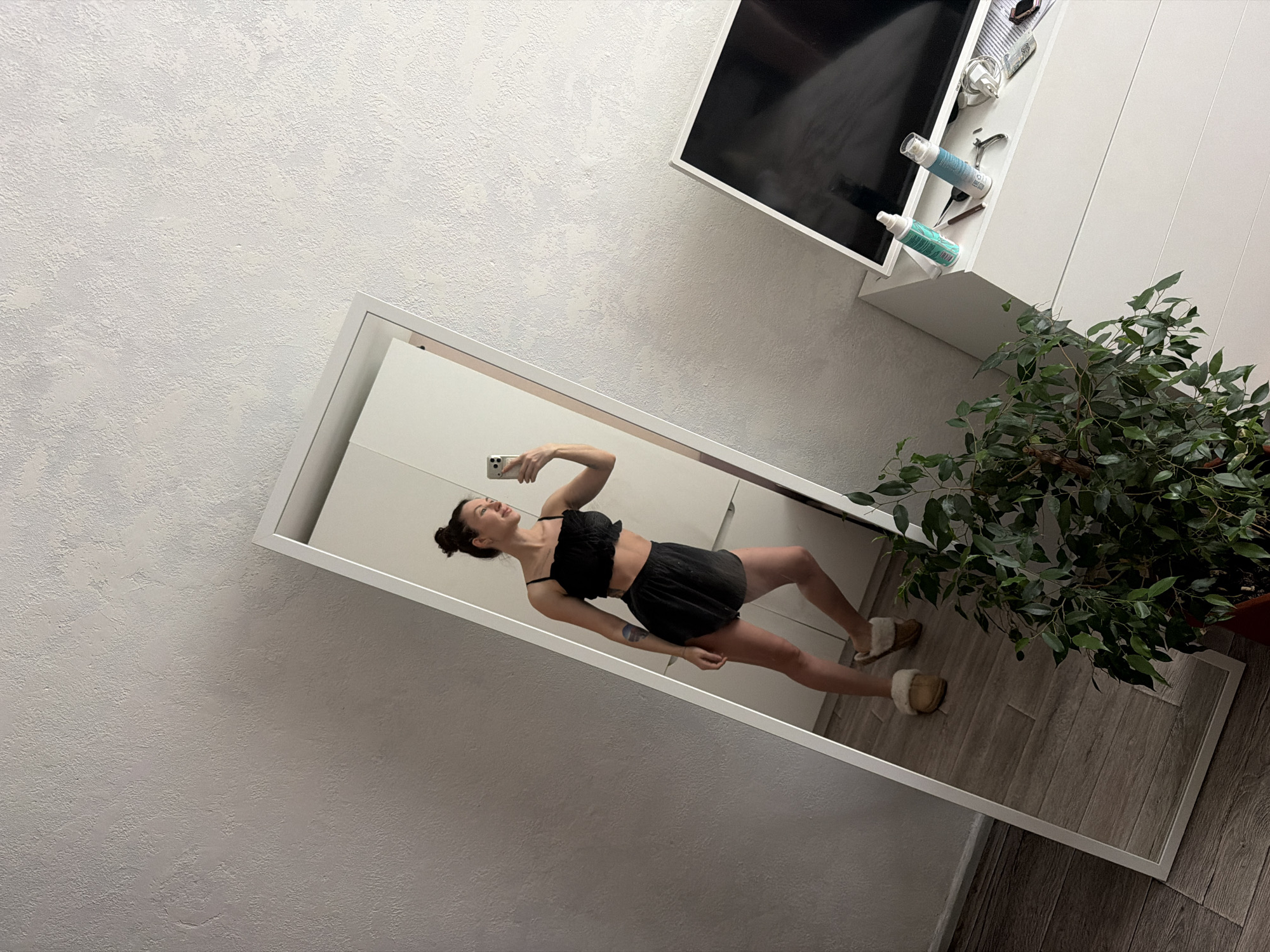 This indoor full-body mirror selfie features a woman in a casual setting in Perm, Russia. The perspective is tilted, with the white-framed mirror angled against a light-colored, textured wall.
The central subject is a woman with dark hair tied in a bun, captured mid-pose while taking the photo. She is wearing a black two-piece outfit consisting of a ruffled crop top with thin straps and matching shorts. Her right arm is raised, holding a light-colored smartphone (likely an iPhone due to visible dual cameras) towards the mirror to capture her reflection. A colorful tattoo is visible on her left forearm. Her expression suggests cheerfulness or playfulness, as she appears to be smiling with her mouth slightly open. She is wearing light brown, fluffy slippers.
To the right of the mirror, a large potted plant with dark green leaves stands on a greyish-brown wood-grain floor. Above the plant and the mirror, a white cabinet or shelf unit is visible. On this shelf, there's a black screen (possibly a television or monitor), two light blue spray bottles, an electrical adapter/charger, and a few other small, indistinct items. The overall lighting is bright and even, suggesting it is daytime. There is no visible text in the image.