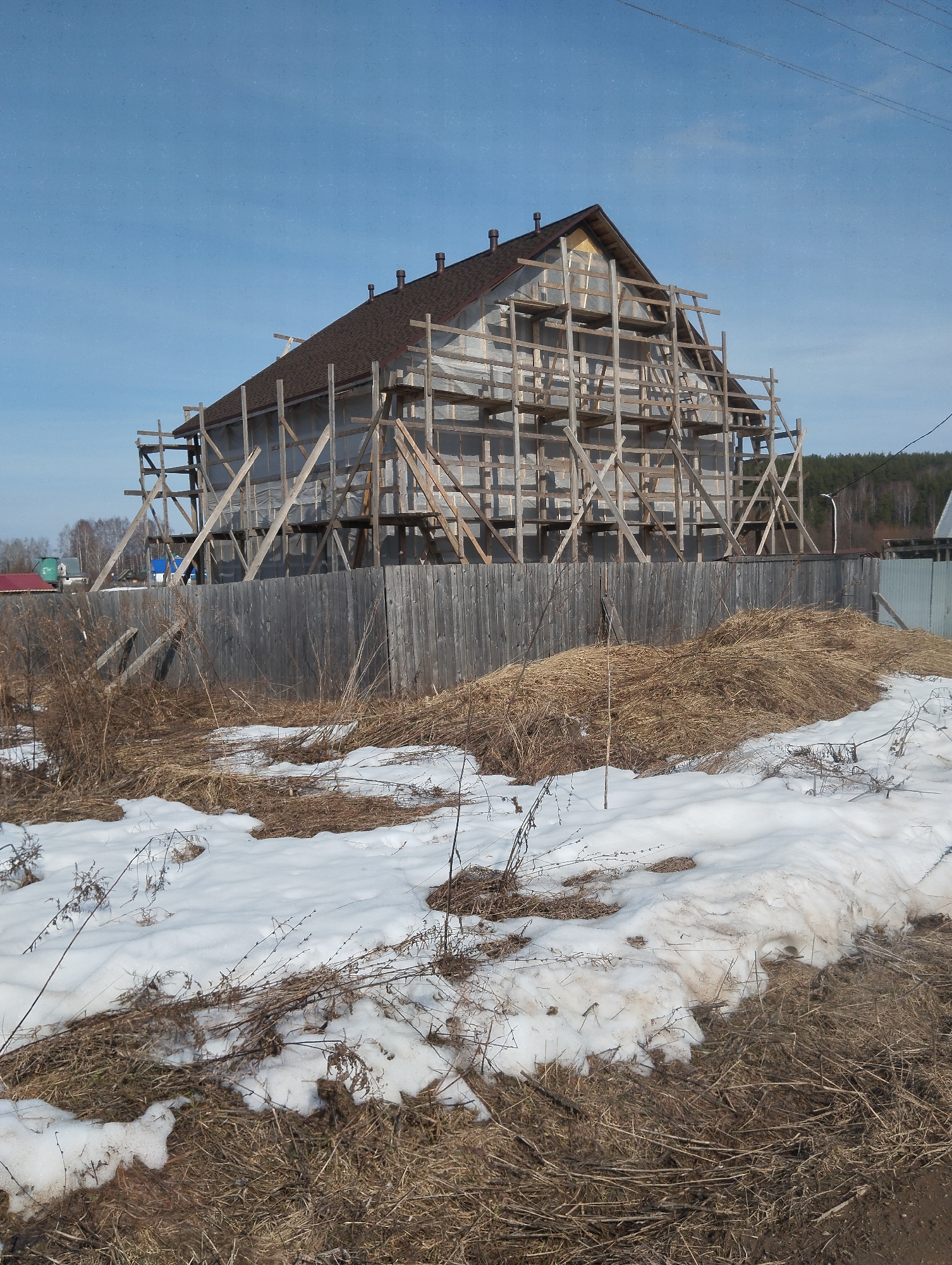 The image shows a house under construction in Grishanki, Russia. The house is surrounded by wooden scaffolding. The roof is covered with brown shingles, and the walls are wrapped in a light-colored material. A wooden fence surrounds the property. In the foreground, there is snow and dry grass. The sky is blue.