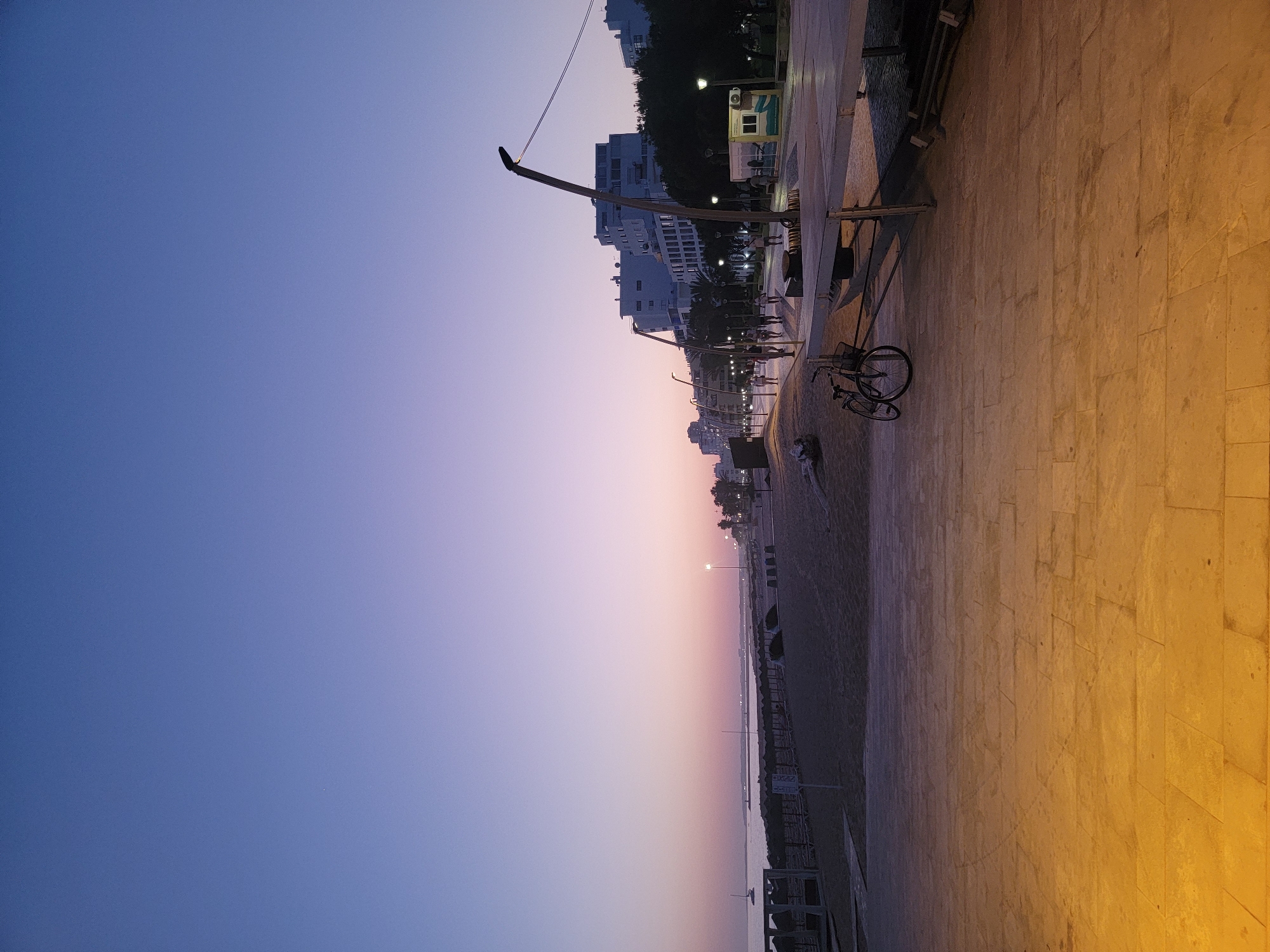 The image captures a dusk scene along the waterfront promenade in Quarteira, Portugal. The sky transitions from a deep blue at the top to pale pink and lavender near the horizon. The sandy beach is visible on the left, with a few thatched sun shelters and loungers set up. Further out to sea, a sailboat is anchored, and a distant lighthouse is visible. The promenade itself is paved with large, rectangular stones that are illuminated by artificial lighting, casting a warm yellow glow. A metal railing separates the promenade from the beach area.
On the promenade, several people are walking and strolling. There are also lampposts lining the walkway, and some trees with dark foliage are present. To the right, a few modern buildings with white facades are visible, suggesting urban development along the coast. In the foreground, a bicycle with a basket is parked. A small, kiosk-like structure is also visible on the right side of the promenade. The overall atmosphere is calm and serene, indicative of early evening in a coastal town.