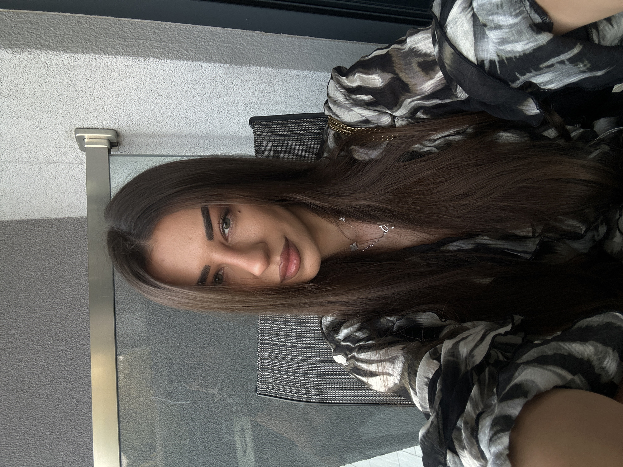 A woman with long, dark brown hair and light green eyes is seated in a chair, looking at the camera. She is wearing a black and white patterned blouse with puffed sleeves. A gold chain necklace with a small pendant is visible around her neck. Behind her, a textured gray wall is visible, with a metal railing and a glass partition. The scene appears to be on a balcony or patio, possibly in a residential area or hotel, given the materials of the wall and railing. There is no specific indication of the location being in Ofrynio Beach, Greece, beyond the prompt.