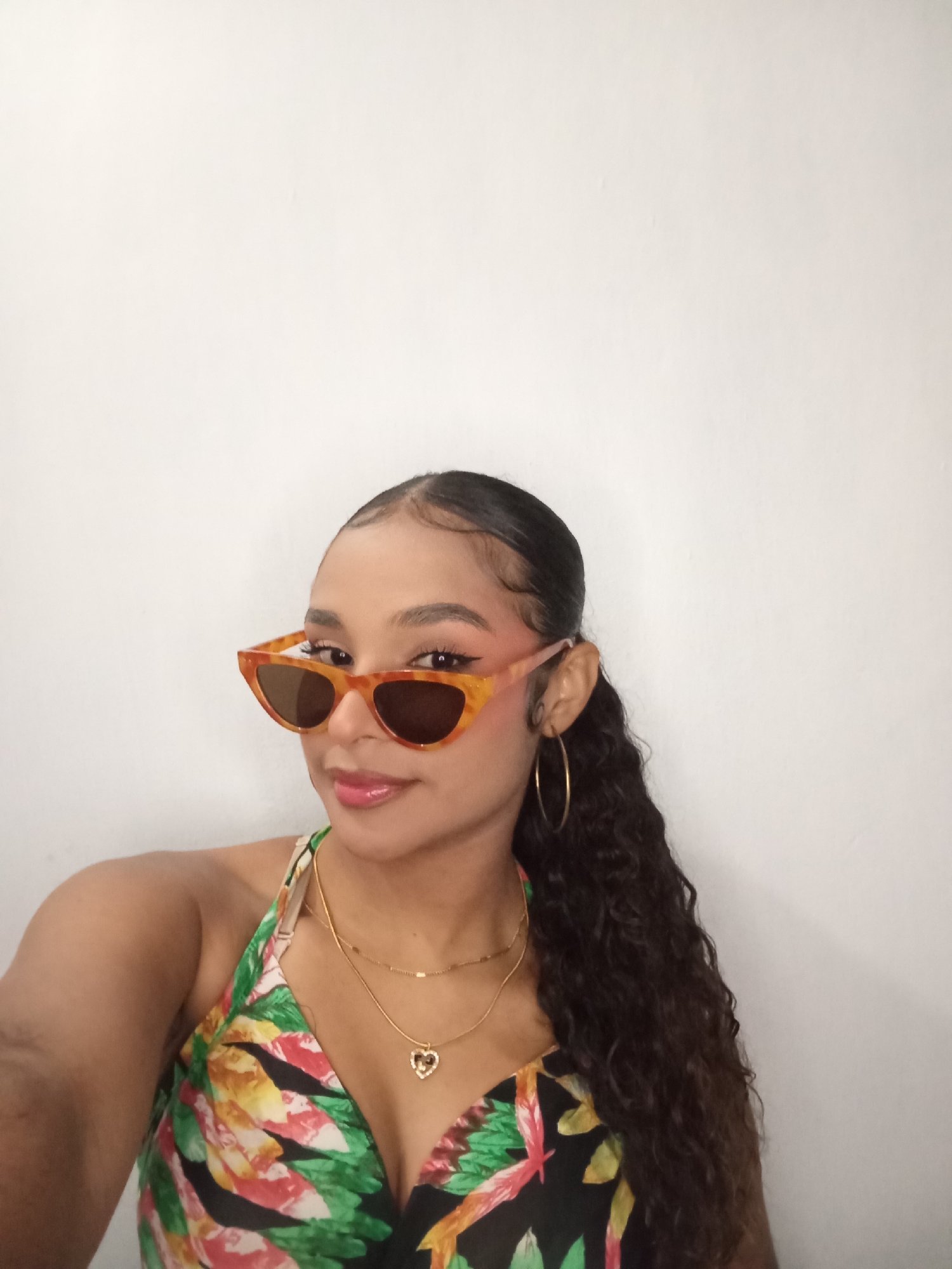 A medium shot depicts a woman with tanned skin, wearing a floral halter top with a plunging neckline. She is adorned with gold jewelry, including hoop earrings and two necklaces, one with a heart pendant. The woman is also wearing orange tortoiseshell sunglasses. Her hair is styled in a sleek, pulled-back ponytail with curly ends. The background is a plain, white wall. No information is available to place this image in Juan Griego, Venezuela.
