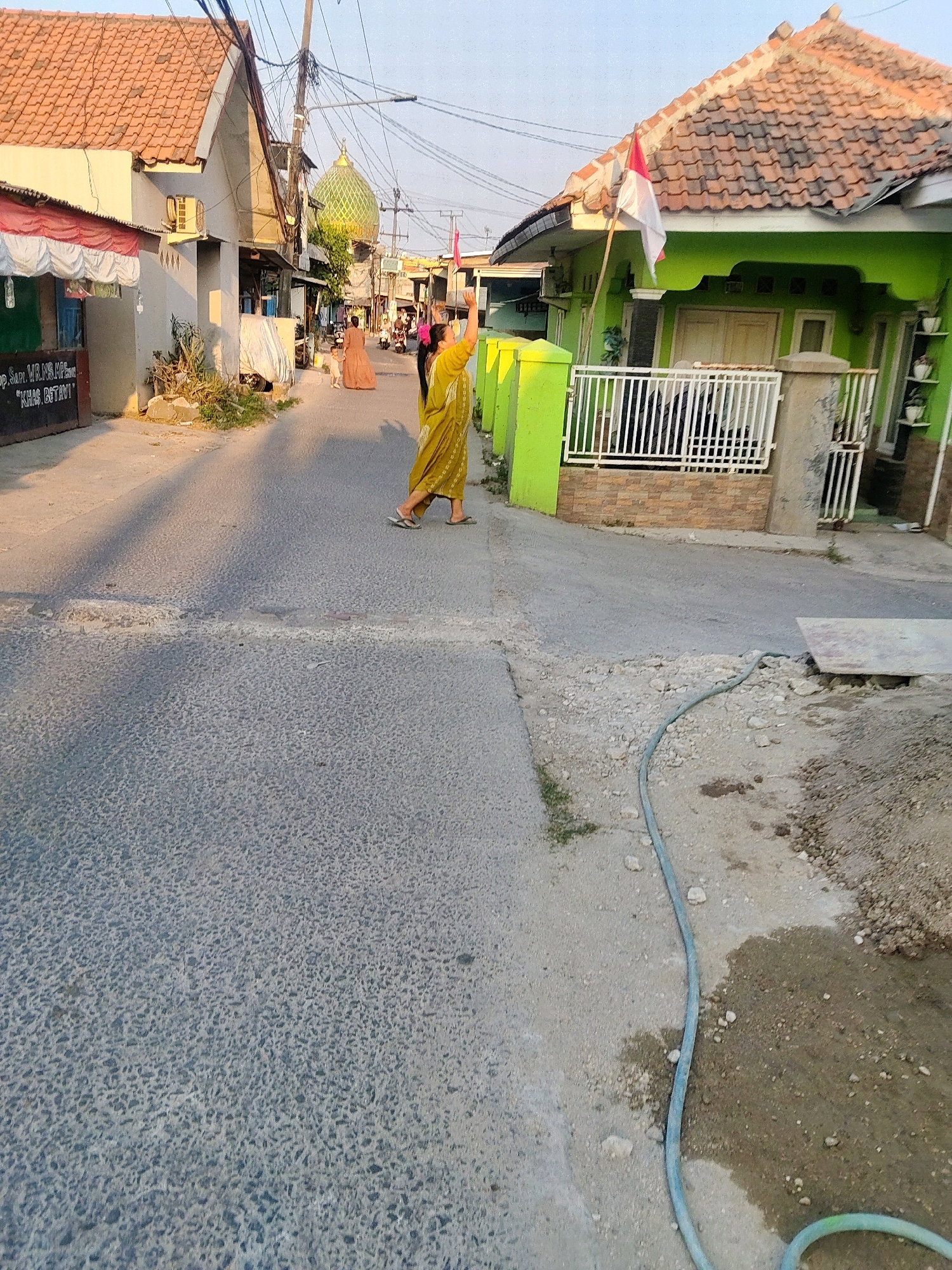 In Mekarmukti, Indonesia, a street scene unfolds under a clear sky. Two women are present. The woman in the foreground, on the right side of the road, is wearing a yellow dress and sandals, with her right arm raised. Another woman in a peach-colored dress is further down the road, walking away from the camera. The street itself is paved with asphalt and shows signs of wear. On the right, a bright green house with a white fence and an Indonesian flag is visible. To the left, a building with a red-tiled roof and white awnings stands. A green dome of a mosque can be seen in the background. The environment suggests a residential area with power lines crisscrossing overhead. A blue hose lies on the ground near the right edge of the frame.