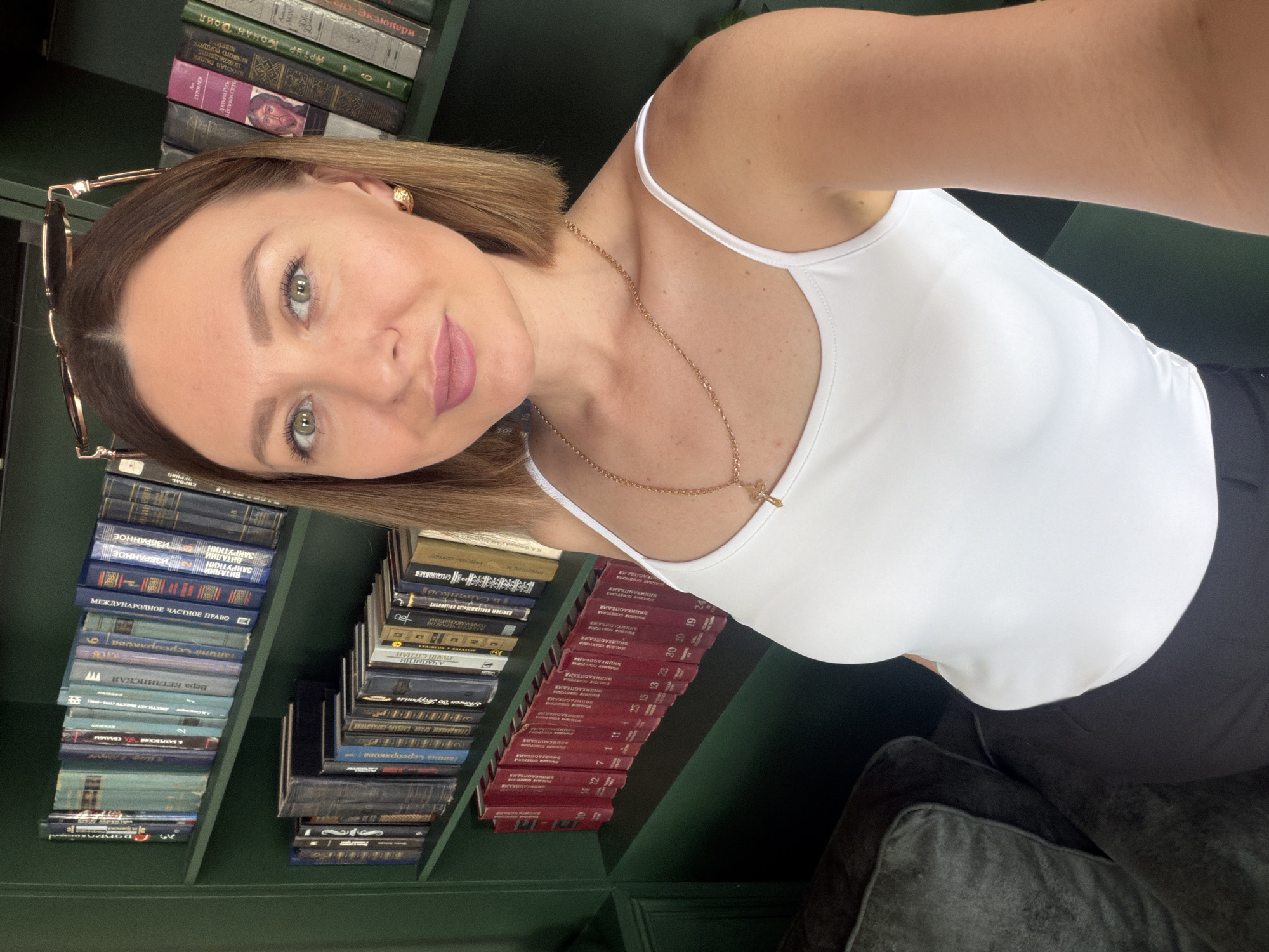A woman with short brown hair is posing for a selfie indoors, in front of a dark green bookshelf filled with books. She is wearing a white tank top and a gold necklace. Glasses are hooked on the bookshelf to the left. The books include Russian titles such as "Encyclopedia," "Vitaliy Zakrutkin," and "Arthur Conan Doyle." The photograph appears to have been taken in Rostov-on-Don, Russia.