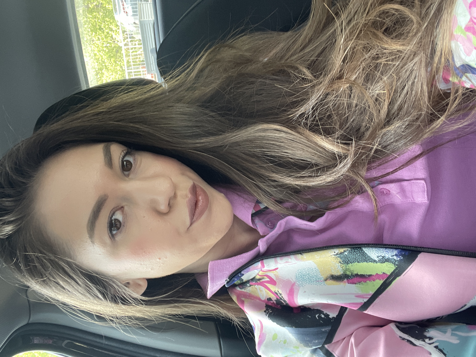 A medium close-up, rotated selfie captures a young woman seated inside a car, likely in Sofia, Bulgaria. Her long, wavy hair, featuring a mix of brown and lighter, golden-blonde highlights, flows over her shoulders and chest. She gazes towards the camera with dark brown eyes, her eyebrows neatly defined, and wearing subtle makeup including eyeliner, mascara, blush, and a glossy, nude-pink lipstick.
She is dressed in a light pink collared polo shirt, over which she wears an open jacket or outer layer. This garment is notable for its vibrant, abstract pattern, incorporating splashes of pink, blue, yellow, and black.
The setting appears to be the interior of a vehicle, with a dark headrest visible behind her. Through a window in the upper left background, lush green foliage and what looks like urban structures are visible, indicating it is daytime. The lighting is bright and natural, illuminating her features and hair. Her expression is calm and pleasant. There is no visible text in the image.