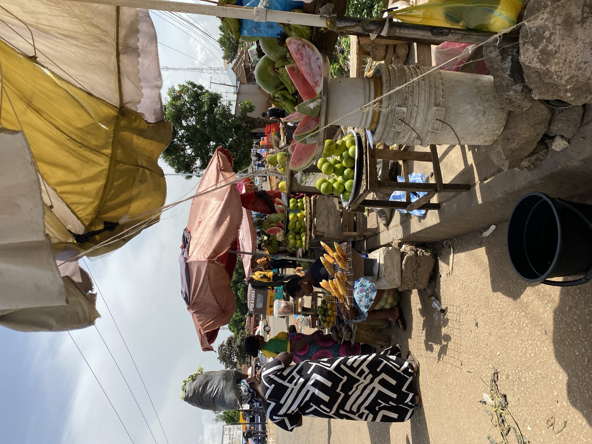 This image depicts an outdoor market scene in Bukuru, Nigeria. The foreground is dominated by a woman in a black and white patterned dress walking towards the viewer. She has a large black bag on her head. To her left, another woman in a colorful dress is interacting with a vendor. Several stalls are visible, laden with various fruits, including watermelons, green apples, and bananas. Corn on the cob is also displayed prominently.
Various objects are present, such as buckets, umbrellas providing shade for the stalls, and what appear to be large stones used for support or display. In the background, more stalls and people are visible, along with trees and buildings. A vehicle with "PTA-10561" on its license plate is parked in the middle ground. The sky is overcast, suggesting a cloudy day. The overall atmosphere appears to be one of everyday commerce and activity.