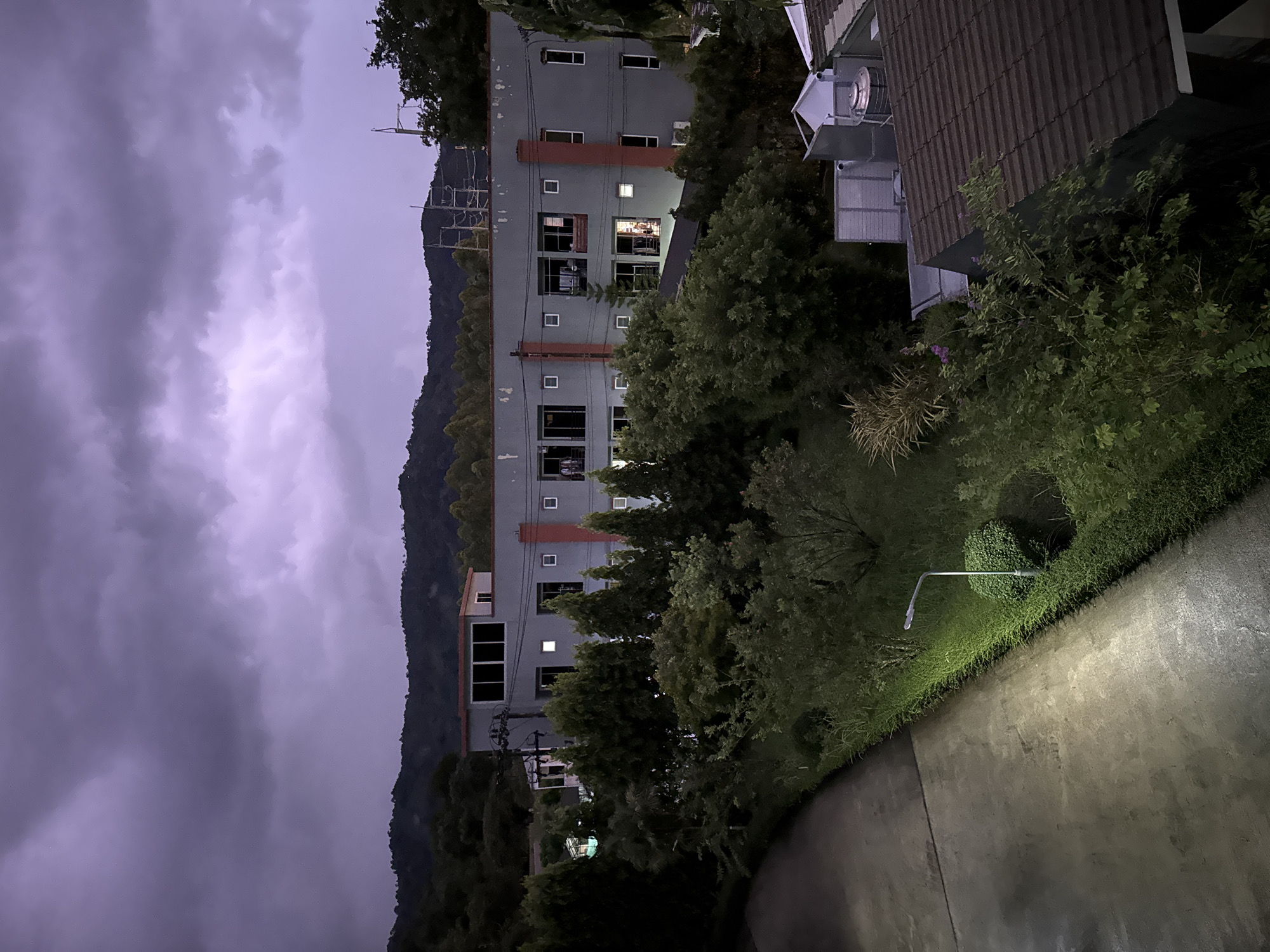The image depicts a nighttime scene in Kathu, Thailand. The dominant features are a multi-story building, a lush green hillside, and a dramatically cloudy sky. The building is painted grey, and it has a series of windows. The hillside behind the building is densely covered with trees and vegetation. The sky is filled with dark, heavy clouds, suggesting an overcast or stormy weather condition. On the right side of the frame, there is a paved area with a lamppost illuminating a patch of grass and bushes. The scene is quiet and appears to be devoid of human activity.