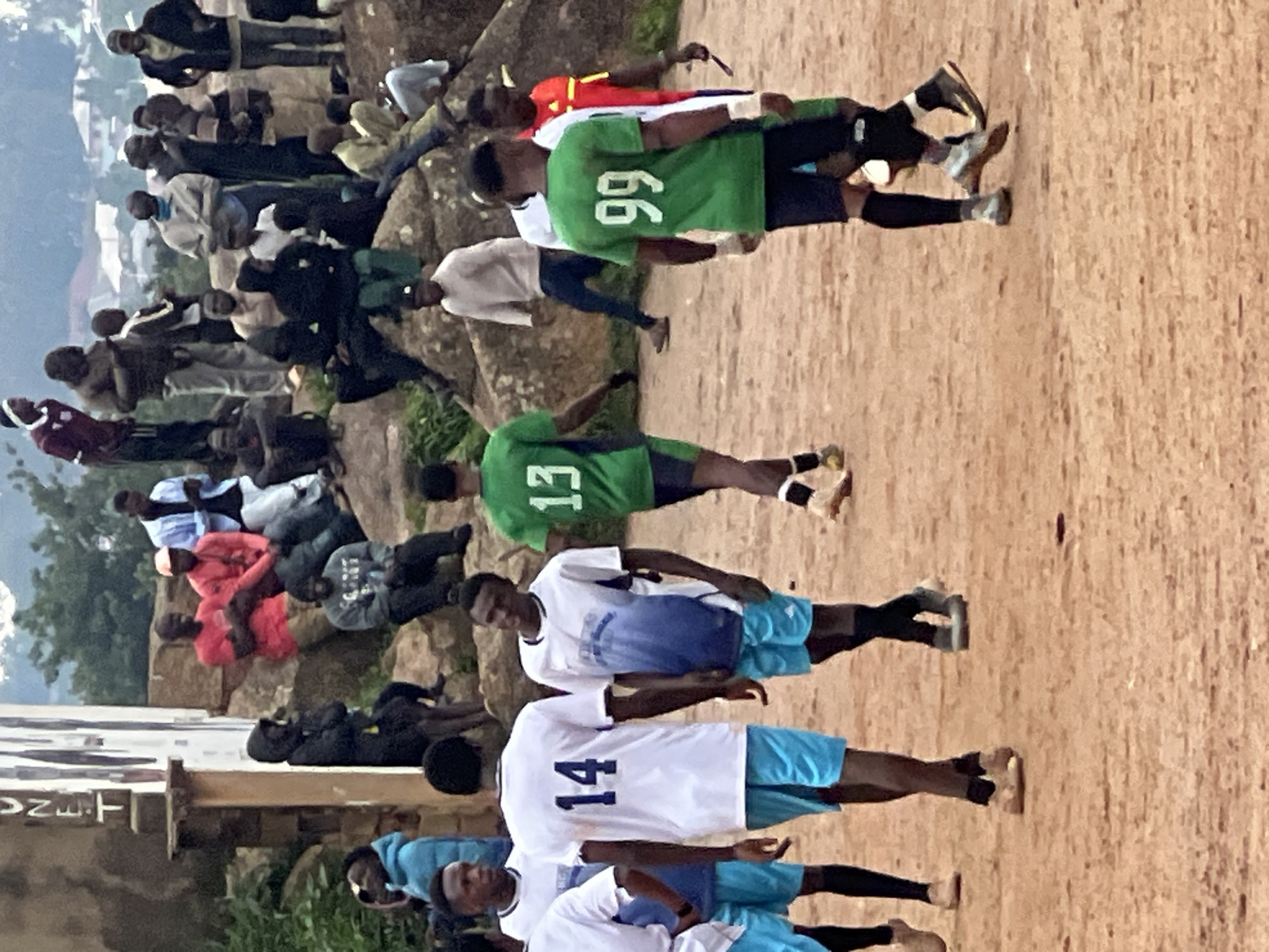 The image depicts a scene outdoors in Jos, Nigeria, likely a public space such as a sports field or a gathering area. Several individuals, predominantly young men, are engaged in or observing what appears to be a soccer game.
In the foreground, four individuals are visible in active or near-active stances, dressed in athletic wear. Two of them are wearing white jerseys with blue accents and blue shorts, one with the number "14" clearly visible on the back of the jersey. Another individual is wearing a green jersey with the number "13" on the back, and a fourth is wearing a green jersey with the number "99". The ground is a reddish-brown dirt or sand, typical of outdoor play areas in Nigeria.
In the middle ground and background, more people are gathered, some sitting on rocks and others standing. They are dressed in a variety of casual clothing. The environment suggests a somewhat rural or undeveloped urban setting, with rocky terrain and vegetation visible in the distance. The presence of a building with lettering, possibly a sign, is noted on the left side of the image. The overall atmosphere appears to be one of a casual community event.