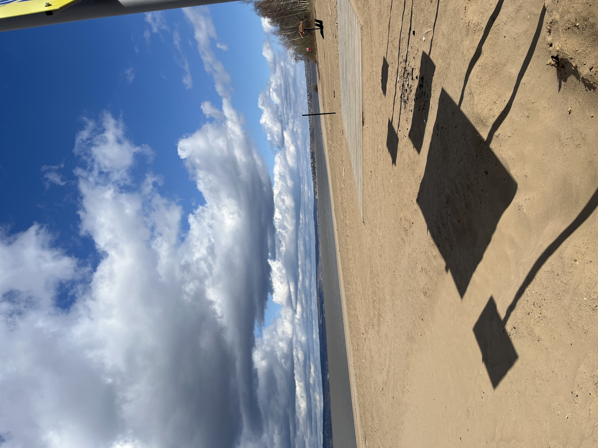 This outdoor image captures a wide-angle view of a sandy shoreline next to a large body of water under a dynamic sky. The scene is likely a beach or riverbank, with the water stretching towards a distant horizon.
In the foreground, a sandy beach occupies the lower and right portions of the frame. Long, distinct shadows are cast across the sand, indicative of a low sun angle (either early morning or late afternoon). These shadows suggest the presence of structures like an umbrella (a large square shadow) and possibly chairs or other equipment (smaller rectangular and elongated shadows). A small pile of debris or ash is visible on the sand where some shadows fall. A light-colored wooden boardwalk or platform extends from the right towards the water. A dark, vertical pole stands in the sand near the boardwalk. At the very top left, a segment of a yellow and gray/silver man-made structure, possibly a canopy or part of some beach equipment, is visible.
The middle ground features a wide expanse of water, likely a river or lake, leading to a distant landmass on the left horizon. This landmass is lined with trees that appear bare, suggesting late winter or early spring. Faint outlines of buildings or a settlement can be seen along the distant shore. A very small, indistinct dark figure, possibly a person or animal, is discernible near the distant tree line.
The sky above is a prominent feature, filled with a mix of bright blue patches and large, fluffy white and gray cumulus clouds, creating a sense of depth and varying light.
There are no clear human activities taking place, and no visible text can be identified. Given the context of Perm, Russia, this could be the Kama River or one of its reservoirs.