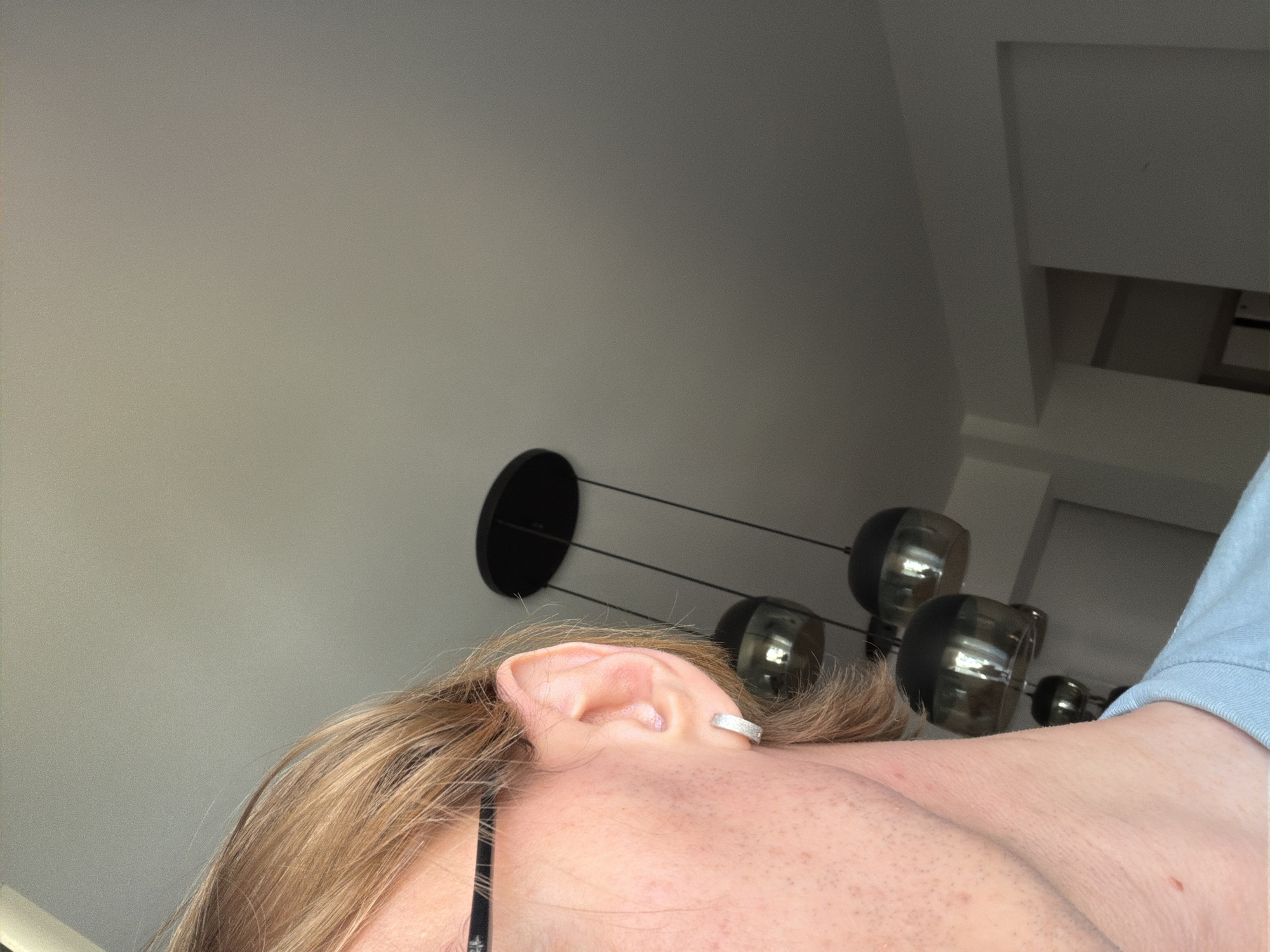 The image depicts an upward-angled close-up view of a person's head and shoulder. Visible are the person's ear, adorned with a silver-colored cuff earring, and light-brown hair. A thin black strap, likely part of clothing or glasses, is also visible. The person is wearing a light-blue top. The background consists of a light-colored wall, a black multi-bulb light fixture, and a glimpse of what appears to be a ceiling and part of a room structure in the upper right corner. The photo appears to have been taken indoors. There is no information in the image to suggest its location in Budva, Montenegro.
