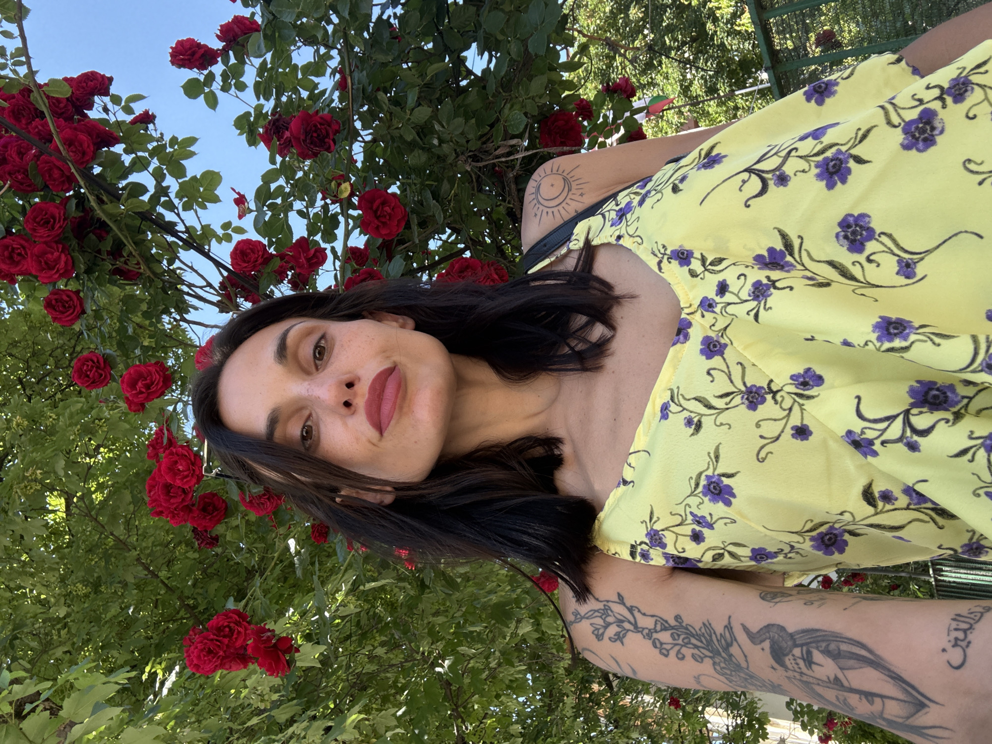 A woman with dark hair stands among red roses under a blue sky. She wears a yellow dress patterned with purple flowers and has tattoos on her arms. The location appears to be outdoors, possibly in a garden or park-like setting, although a precise location in Pleven, Bulgaria, cannot be determined from the image.