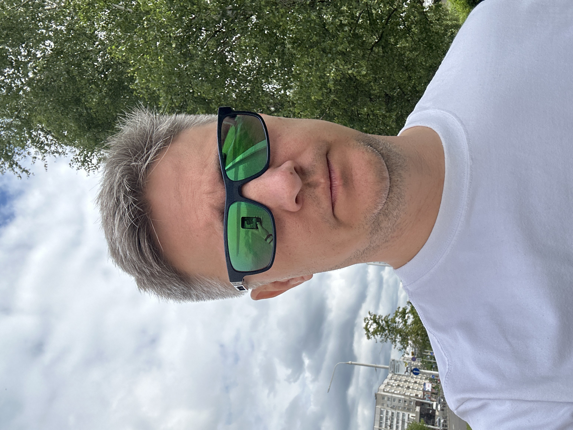 The image, oriented sideways, shows a man outdoors in what is contextually Minsk, Belarus. The man is depicted from the chest up, facing the camera with a neutral expression. He has short, graying hair and is wearing a white crew-neck t-shirt. Dominating his face are green-mirrored sunglasses with black frames. In the reflection of his left sunglass lens, a hand holding a smartphone, presumably taking the picture, is visible.
The setting appears to be a city environment on an overcast day. Above the man, lush green tree foliage is visible, indicating proximity to nature. Below him, the cloudy sky fills a significant portion of the background, with glimpses of distant urban structures, including a multi-story building and a street light, suggesting a street or park area within the city. No discernible text is visible on any objects or in the background.