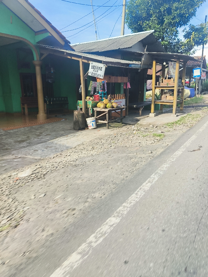 The image shows a street side view of a small shop in Sukatani, Indonesia, located at latitude: -6.643, longitude: 107.415. The shop is situated on the right side of the road. It has a wooden structure with a sign that reads "SEDIAMIE AIR KELAPA" (providing coconut water). The shop is selling coconuts, and there is a white plastic bucket, a large metal drum, and a wooden table with coconuts. The shop is facing a paved road, with a white dividing line. The road is dusty and there are no cars or people visible. The shop is located in front of a house on the left side of the image. The house has a green facade and a covered porch with a single column. The background of the image is a blurred green landscape.