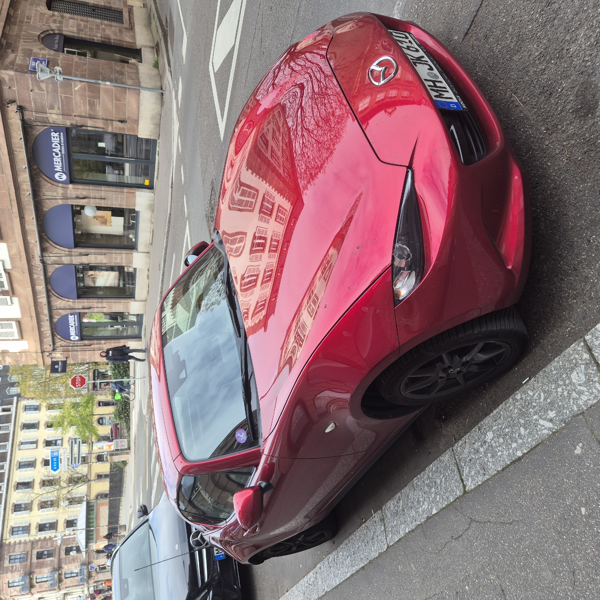 The image captures a street scene in Strasbourg, France, featuring a red Mazda MX-5 (Miata) parked along the side of a street. The car has a German license plate, which reads "MHJK 620". The buildings across the street have signage for businesses, including "MERCADIER Teintes & Matières" and "FARROWA BALL". The buildings are made of red brick with light-colored windows. A stop sign is visible on the left. People are walking and riding bicycles in the background. The car's hood has reflections of the surrounding buildings.