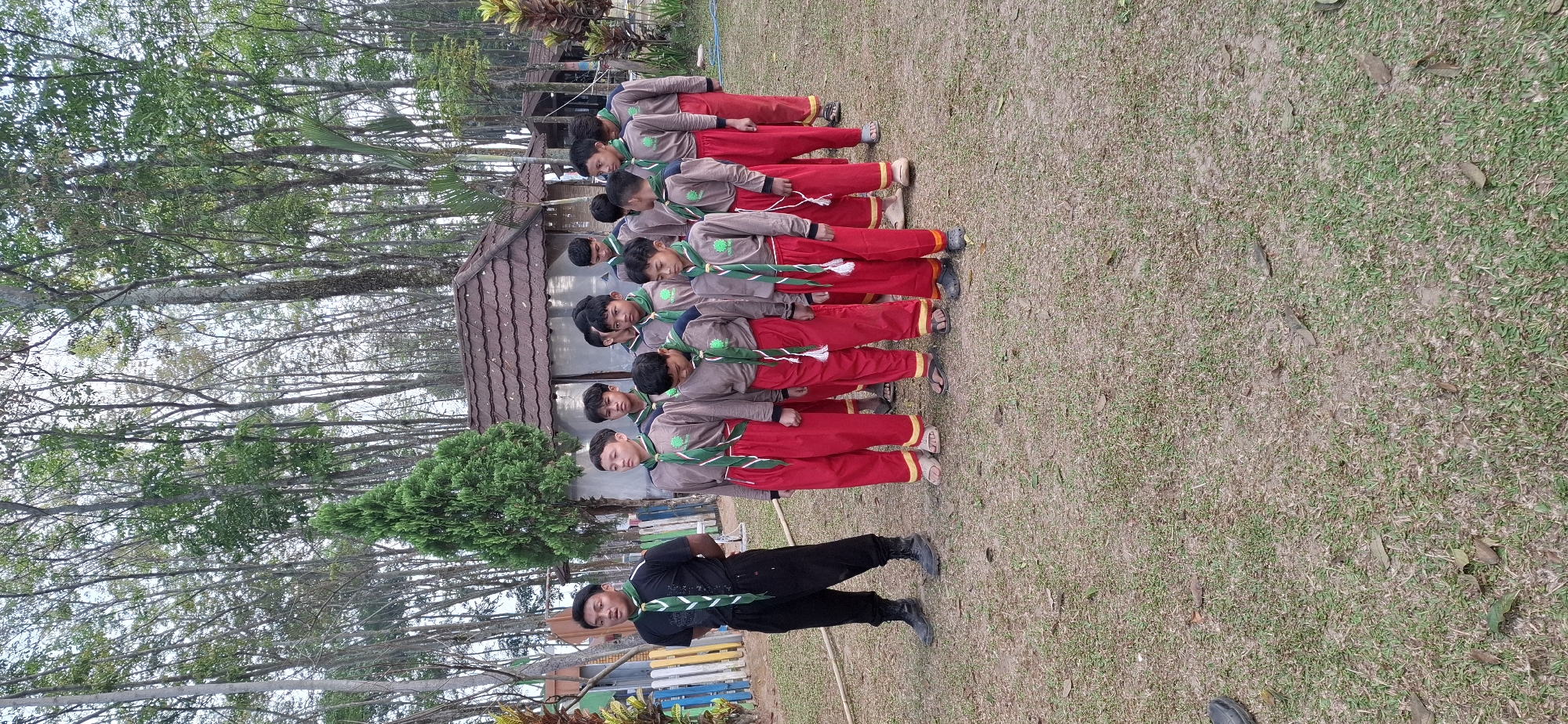 A group of young boys in scout uniforms stand in a line on a grassy area in front of a small building with a brown roof. The boys are wearing red pants and light brown shirts with green scarves. One boy in the front left is wearing a black shirt and black pants, also with a green scarf. The background consists of numerous tall trees with thin trunks and green leaves, suggesting an outdoor setting in a wooded or park-like area. The building appears to be a simple structure with a large window on the front. There are colorful wooden posts to the left of the group of boys. The ground is covered in dry grass with some fallen leaves. The lighting suggests it is daytime, possibly overcast or in a shaded area. The location is identified as Puntukrejo, Indonesia.