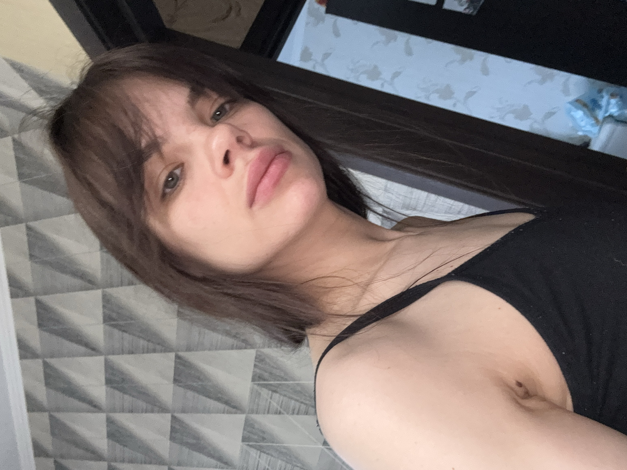 The media file is a selfie of a young woman, likely taken in Rostov-on-Don, Russia. She has long, dark brown hair with bangs covering her forehead and light-colored eyes, which appear green or grey. Her lips are full, and her expression is neutral, appearing somewhat serious or pensive. She is wearing a simple black tank top or camisole with thin straps. The image is tilted, suggesting she might be reclining or taking the photo from an overhead angle.
The background shows an indoor setting. To the left, there is a wall covered with modern, three-dimensional gray and white geometric tiles, forming a repeating pyramid-like pattern. A dark wooden or metal beam or door frame runs horizontally across the upper part of the frame, separating the tiled wall from another wall in the distance. This distant wall, visible on the right, features lighter, possibly white or light blue, wallpaper with a subtle, faded floral or abstract design. A small, indistinct blue object, possibly a plastic bag, is partly visible in the upper right corner against this patterned wallpaper. There is no visible text in the image.