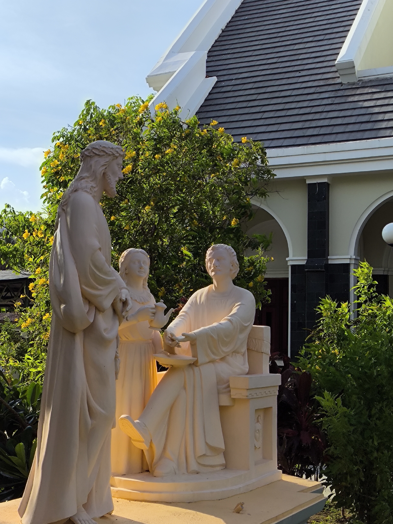 A religious sculpture depicts Jesus, a young woman, and a man. Jesus stands to the left, looking down towards the other two figures. The young woman, holding a jug and cup, stands in the center, slightly to the right of Jesus. To the right, a man is seated on a stone bench, looking at Jesus. The sculpture is made of a light-colored material and is placed outdoors. Behind the sculpture is a building with a dark tiled roof and arched entrances. Lush green trees with yellow flowers are visible behind and to the sides of the sculpture, with some dark red foliage in the foreground to the right. The scene appears to be set in a courtyard or garden area of a religious establishment. The image was taken in Bac Nha Trang Ward, Vietnam.