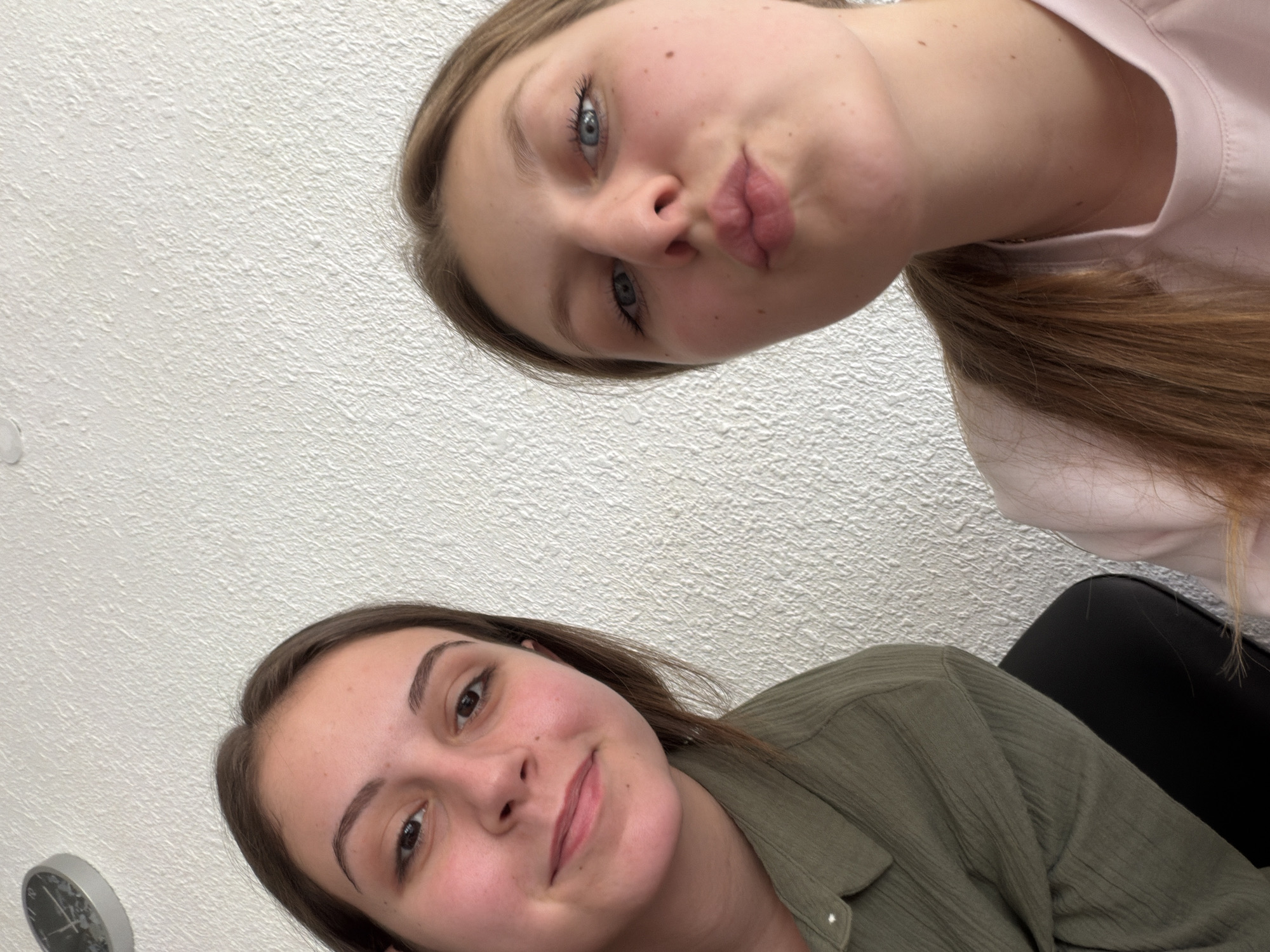 The image captures a selfie taken by two young women indoors, likely in a home environment. The wall behind them has a textured white finish. The woman on the left is looking towards the camera with a slight smile, wearing a green collared shirt. The woman on the right is puckering her lips as if for a kiss, with her eyes looking directly at the camera. She is wearing a light pink shirt. A portion of a round clock can be seen in the upper left corner of the image. There is no discernible information in the image to suggest a location in Burgas, Bulgaria, or any urban environment.
