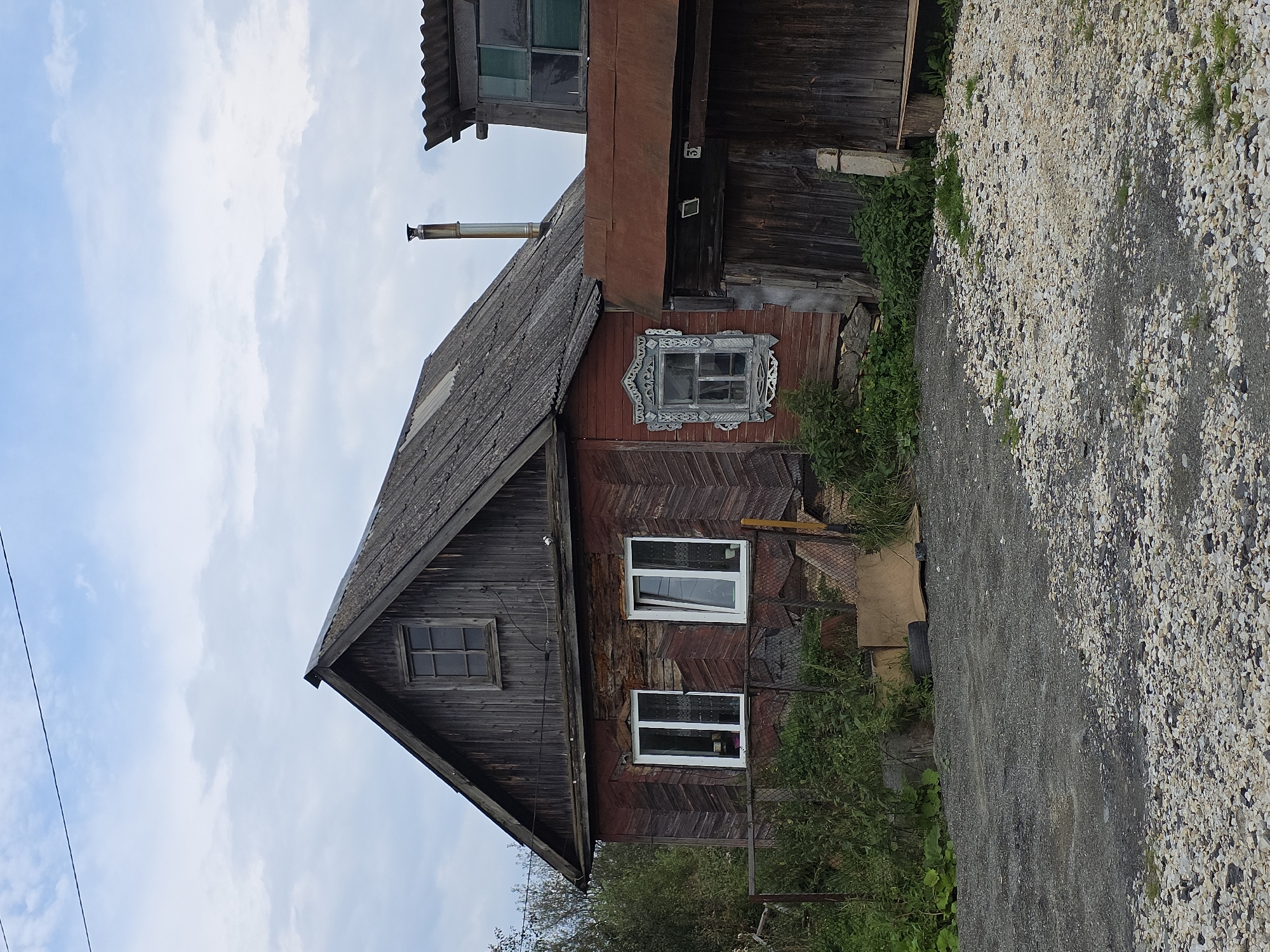 The image depicts a rustic, weathered wooden house in a rural setting, likely in Magnitka, Russia. The house has a prominent gabled roof covered in corrugated material, and the exterior walls are constructed of aged, red-painted wooden planks arranged in a chevron pattern. Two white-framed windows are visible on the main floor, one of which is slightly ajar. Above these, a smaller, square window with a grid pattern is set into the upper gable. A decorative, blue-painted wooden frame surrounds a single window on the side of the house. Adjacent to the main house, a separate structure with a corrugated metal roof and wooden walls is partially visible. A metal chimney pipe extends vertically from the roof of the main house. The foreground is covered with a mixture of gravel and dirt, with overgrown green vegetation encroaching from the sides. A wire fence runs along the front of the property. The sky above is overcast with scattered clouds.