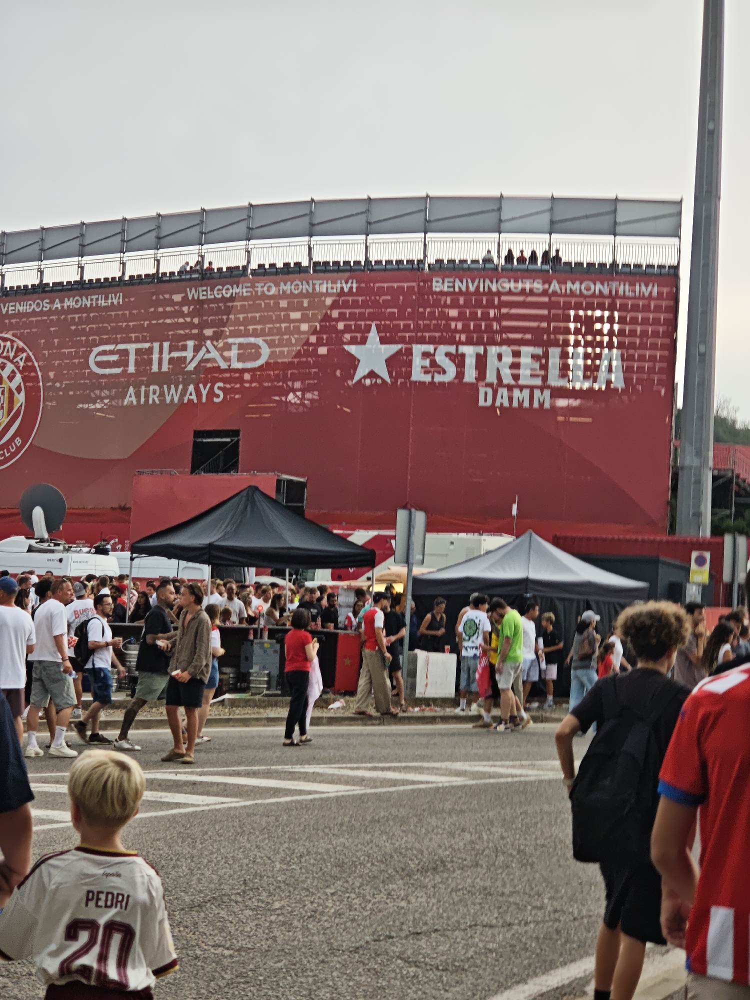 The image depicts an outdoor scene, likely at a stadium or event venue, with a large red building in the background featuring advertisements for "ETIHAD AIRWAYS" and "ESTRELLA DAMM." The building has "VENIDOS A MONTILIVI" and "WELCOME TO MONTILIVI" text. Numerous people are gathered in front of the structure and walking on a paved area. Some individuals are standing in small groups, while others are in motion. There are tents set up, and in the foreground, a young boy wearing a white and maroon jersey with the name "PEDRI" and the number "20" is facing away from the camera. The overall atmosphere suggests a public gathering or event.