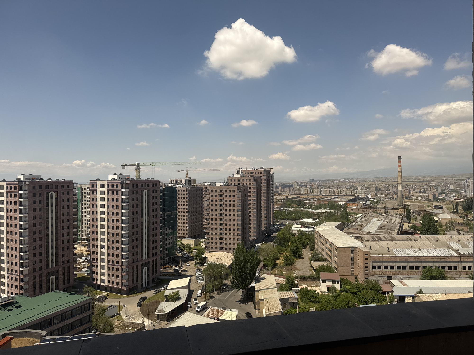 An aerial, eye-level, wide shot captures an urban landscape under a clear blue sky with scattered white clouds. In the foreground, a dark railing runs across the bottom of the frame. The middle ground is dominated by several tall, multi-story residential buildings with reddish-brown exteriors and numerous windows. One building in the left foreground has a green, slanted roof. To the right of these apartment buildings, an industrial area features a long, low-rise building with a corrugated metal roof and an adjacent tall smokestack with red and white stripes. Construction cranes are visible in the distance, suggesting ongoing development. The background shows a sprawling city with more buildings and a hazy horizon. Several cars are parked along a road winding through the scene, and some trees are scattered throughout the urban and industrial areas.