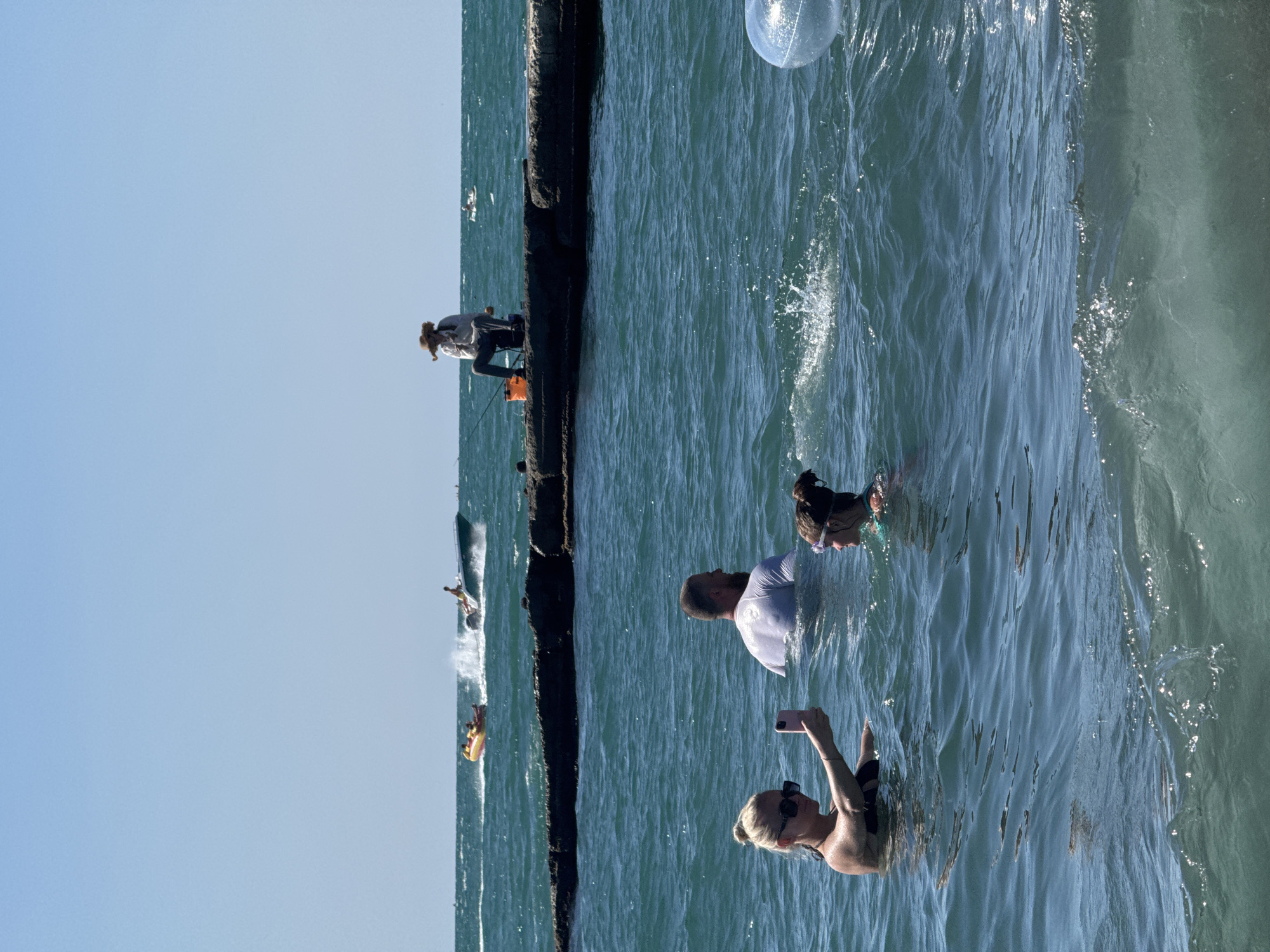 In Sochi, Russia, a sunny day unfolds at the sea. In the foreground, a woman in a black bikini takes a selfie with her pink phone while standing in the water. Beside her, a younger person wearing goggles looks to the right. A man with a beard, wearing a white t-shirt, is also in the water behind them. Further out, a person in a straw hat sits on a rocky breakwater, fishing with a rod and an orange bucket. In the background, a motorboat pulls an inflatable tube with people riding on it. A larger motorboat speeds across the water, creating a wake. The sky is clear blue, and the sea is choppy. A large, clear inflatable sphere is partially visible in the lower right corner of the image.